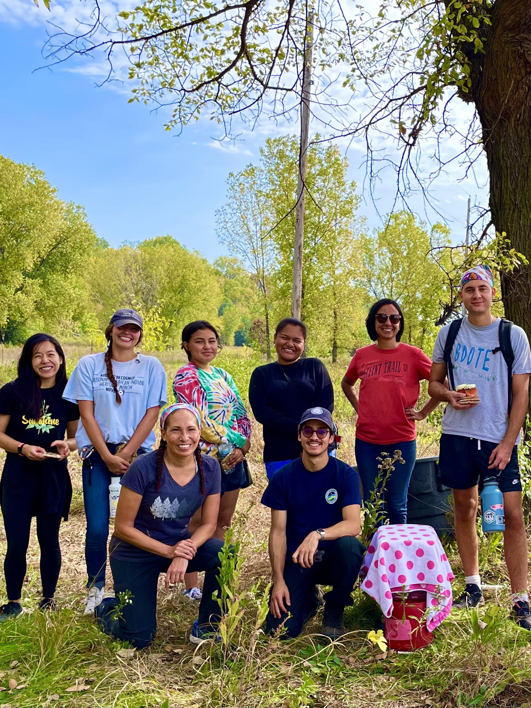  Mishaila Bowman - Lower Phalen Creek Project (second from left)  A group of people stand together to pose for a picture under a tree near a prairie. It is a bright sunny day and everyone is smiling. 
