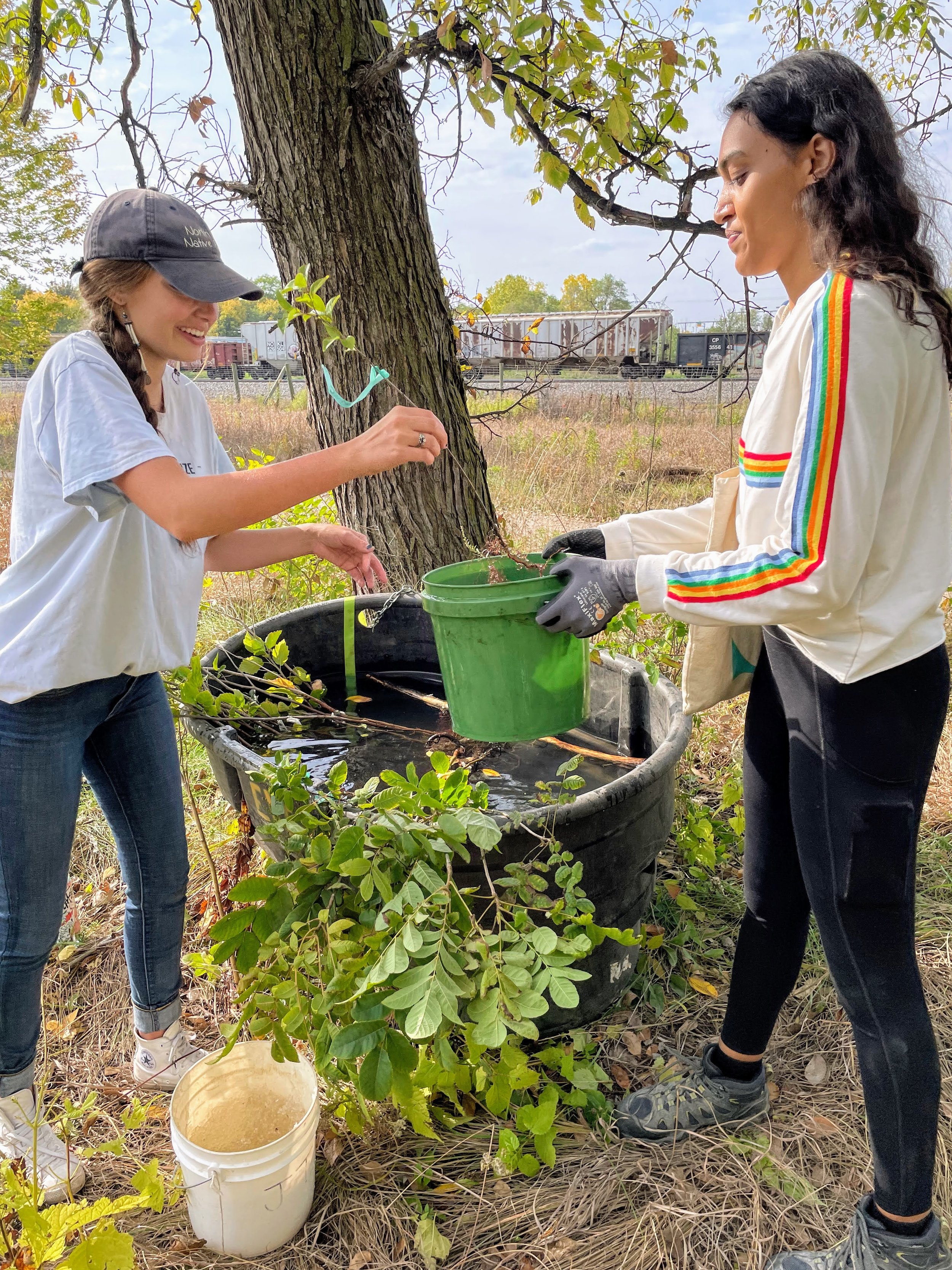  Mishaila Bowman - Lower Phalen Creek Project (left)  A person holds a plant baby in their hand and another person holds a bucket of water. They are both standing next to a tree near a prairie. 