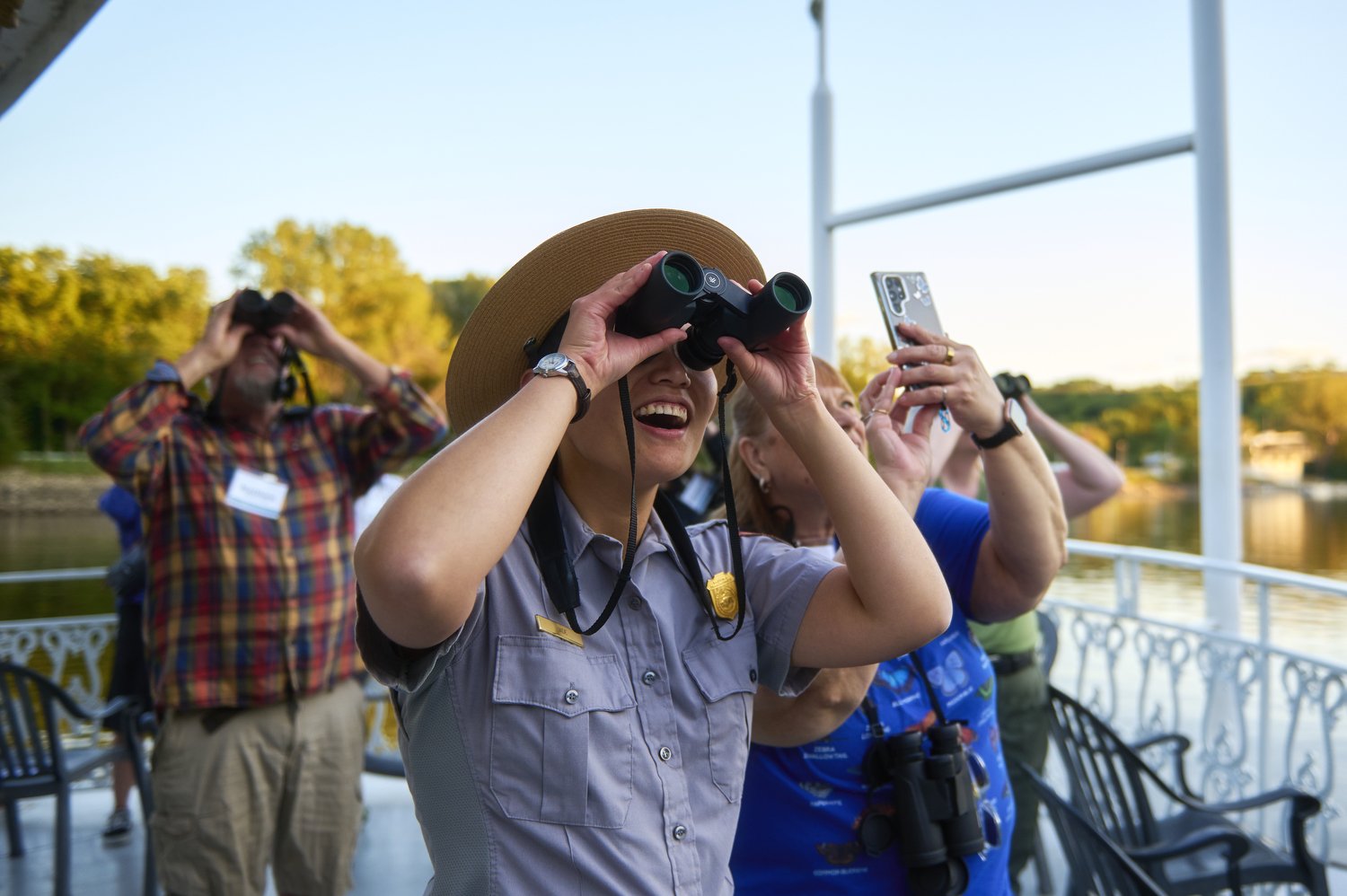 Park ranger and others using binoculars and cameras to look into the sky to see wildlife while on a paddle boat. The white railing of the boat and the river and greenery of the far bank are in the background.