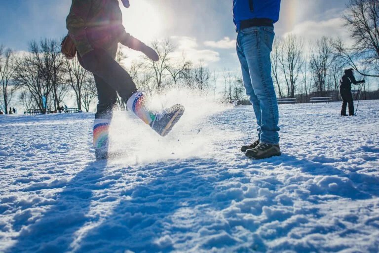 Snowy landscape with trees in the background and sun and two people in the foreground (we only see the bottom 2/3 of them) in winter gear, kicking snow.