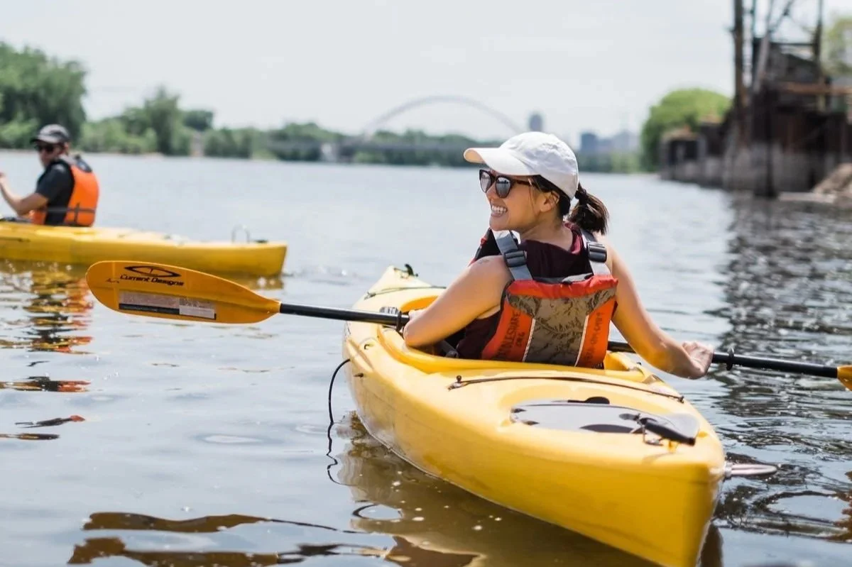 Person in a yellow kayak with a white hate and orange personal floatation device holding a paddle while floating on the river. Ahead is another paddler, greenery, and urban sites in the park.