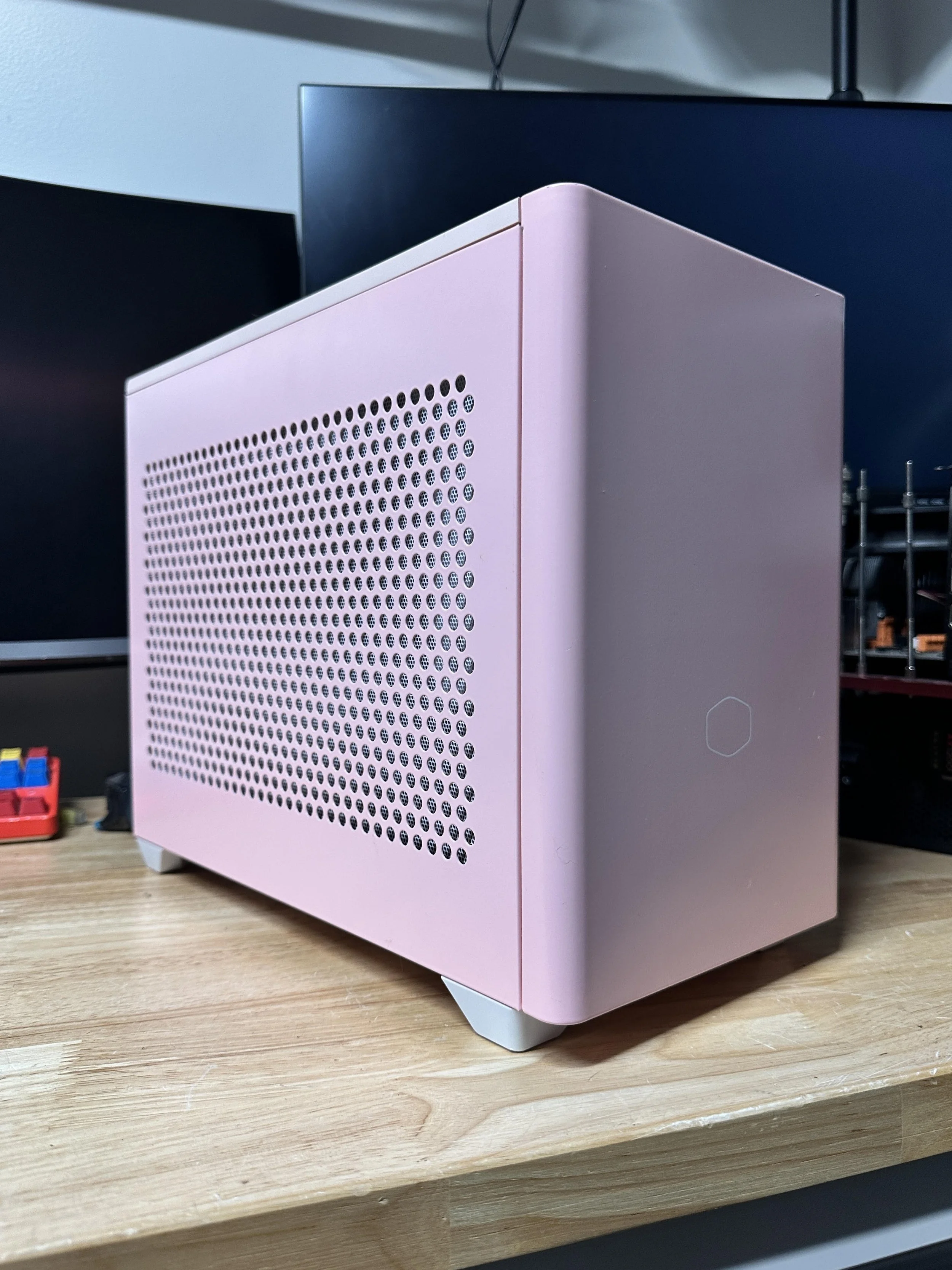 A pink computer case with vent holes on a wooden desk, with a black monitor and a small container with colored objects in the background.