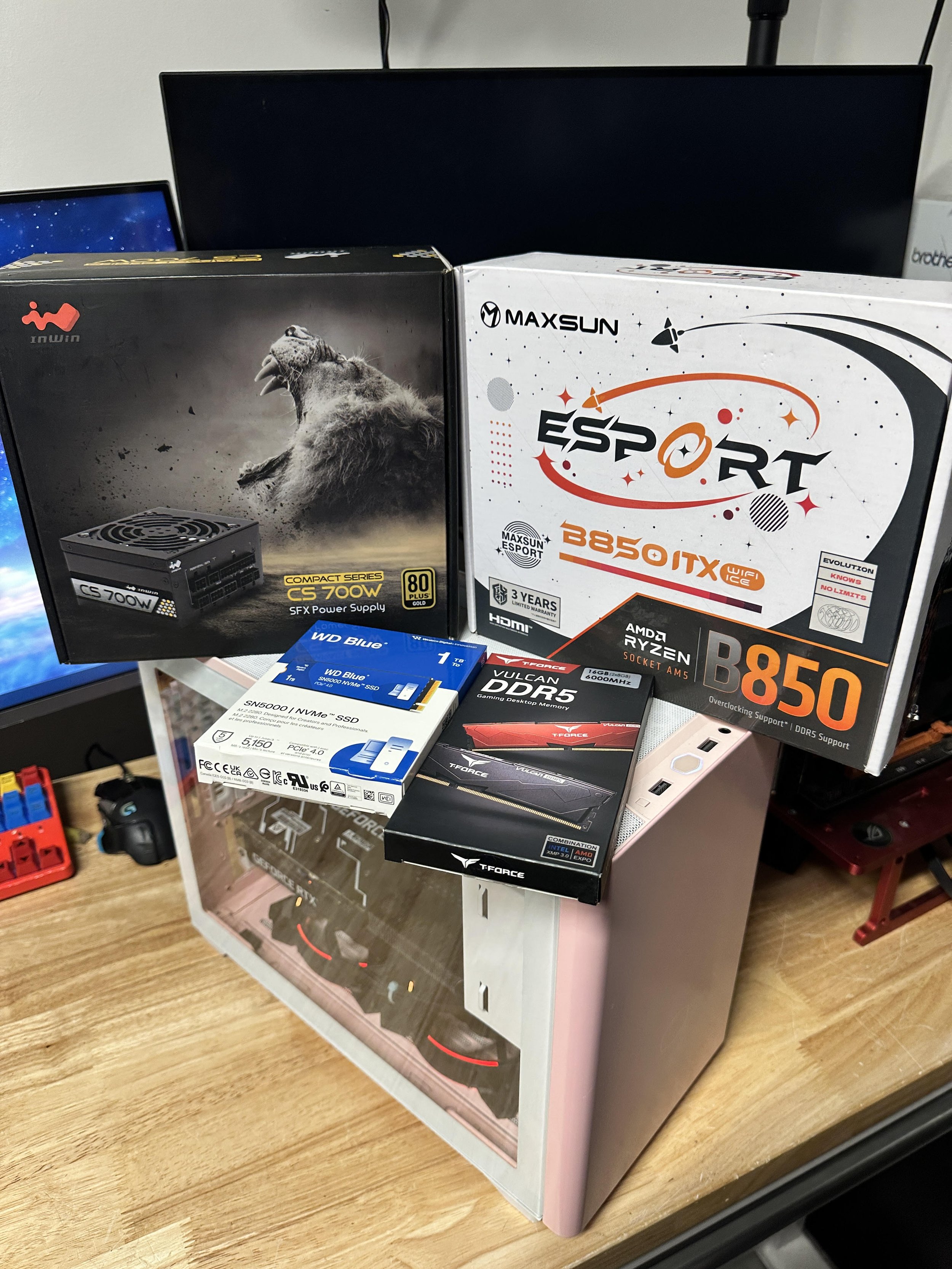 Unopened computer components including a power supply, motherboard, SSD, RAM, and a prebuilt gaming PC case on a wooden desk.