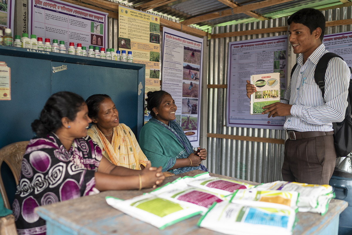 Kalpana and other farmers sit inside a small agro-input shop and listen to a representative from ACI, an agri-input company working across Bangladesh.