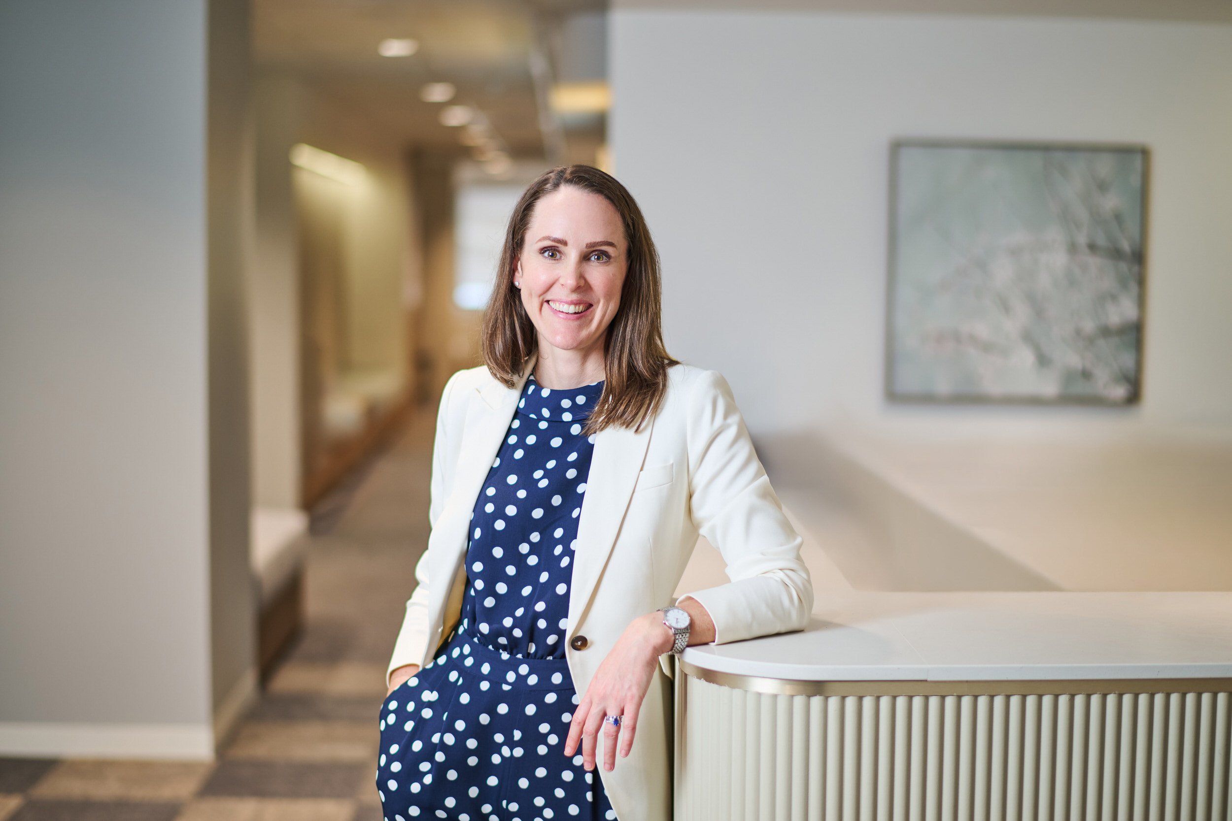 A hearth industry lifestyle corporate headshot of a woman with shoulder-length brown hair, wearing a white blazer over a navy blue dress with white polka dots, standing and smiling in an indoor setting with neutral-colored walls and artwork.