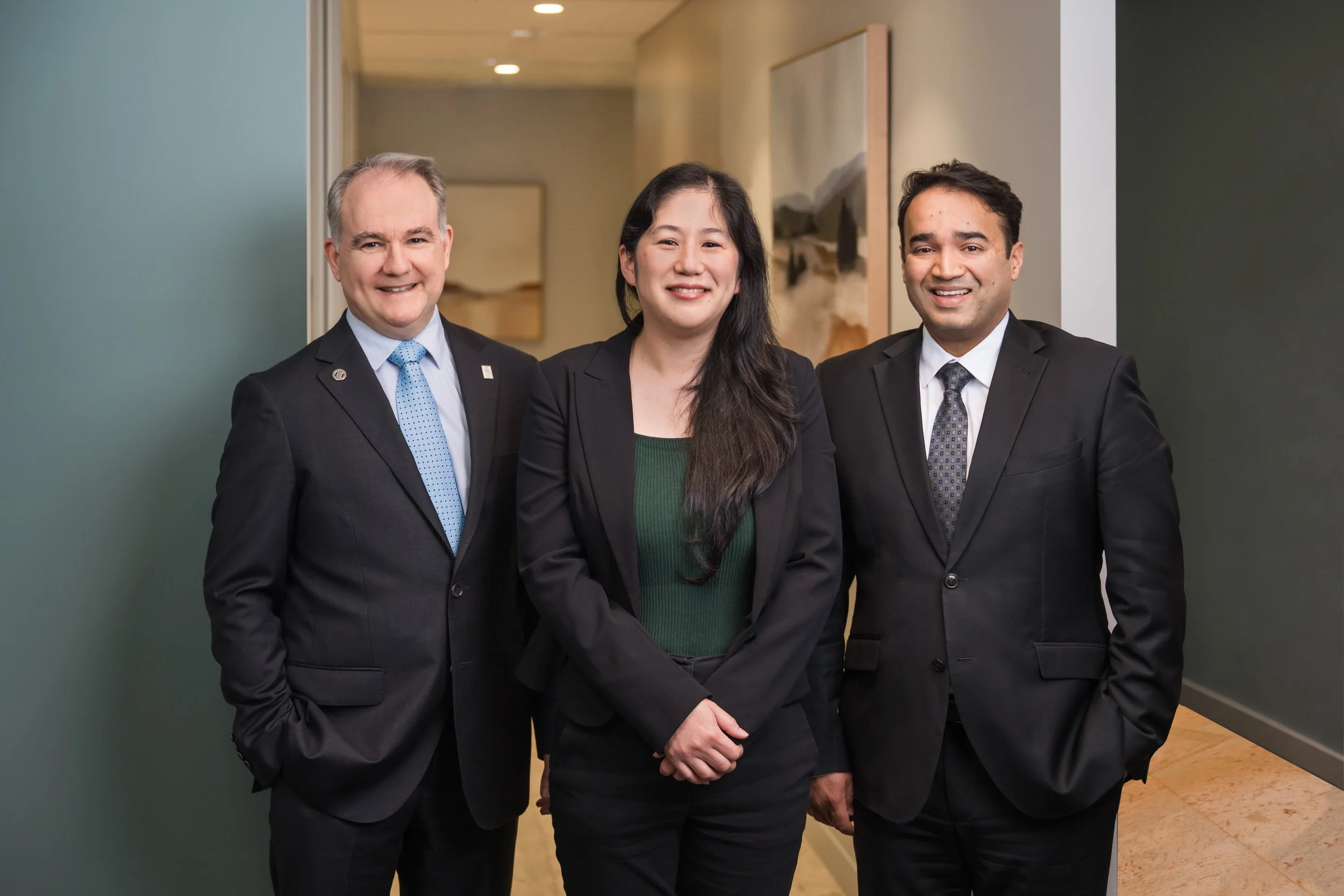 Classic corporate team photography of three professionally dressed individuals standing indoors, smiling for the camera, with artwork and wall decor in the background.