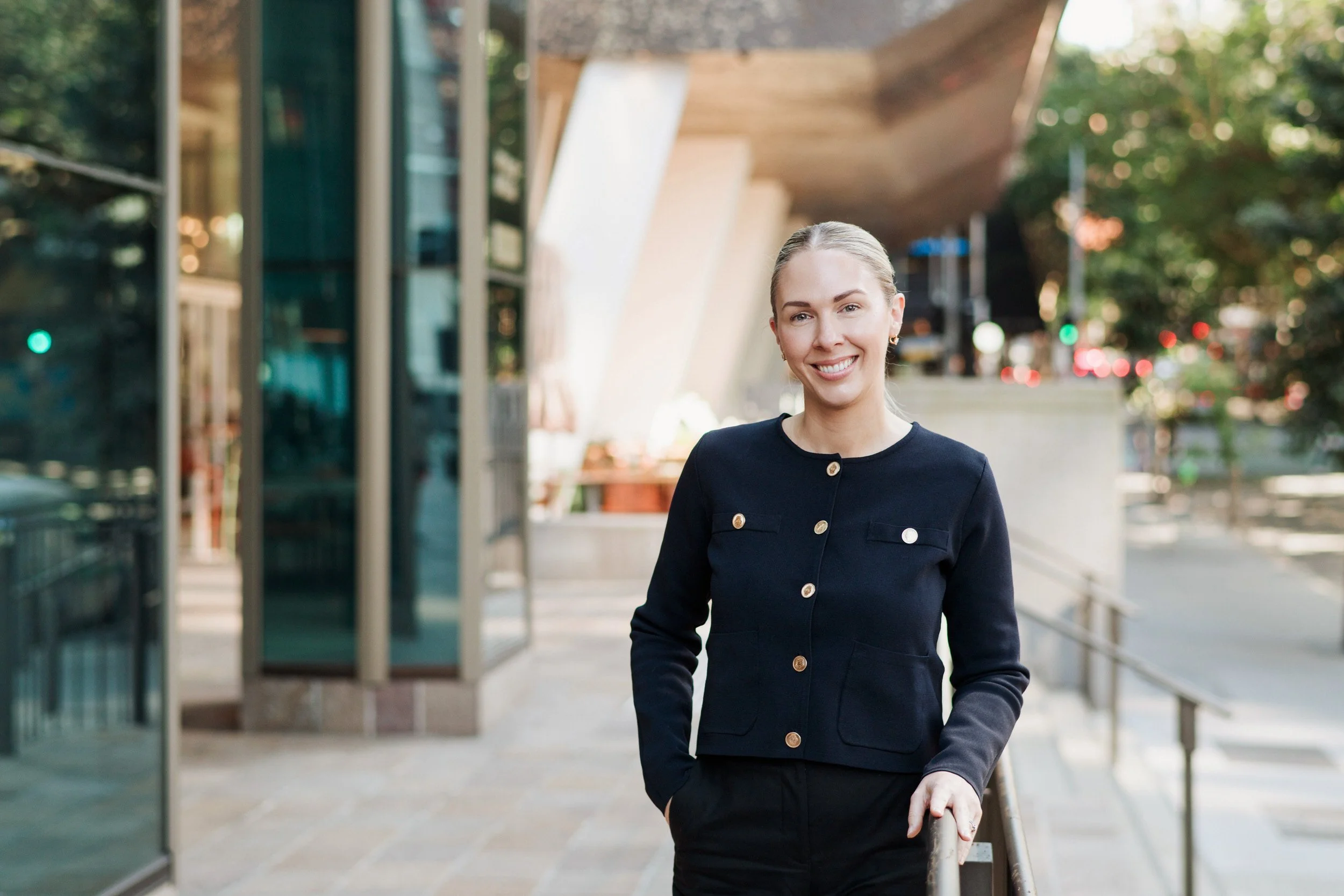 A woman with blonde hair pulled back, wearing a black blazer with gold buttons, smiling while walking outside on a city sidewalk next to a glass building with greenery and trees in the background.
