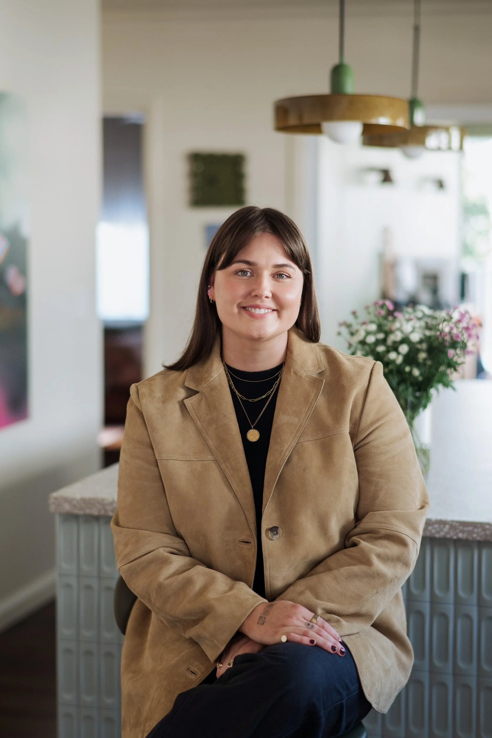 A young woman with shoulder-length brown hair, wearing a tan blazer and black shirt, sitting in a well-lit room with a large window and flowers in the background, smiling at the camera.