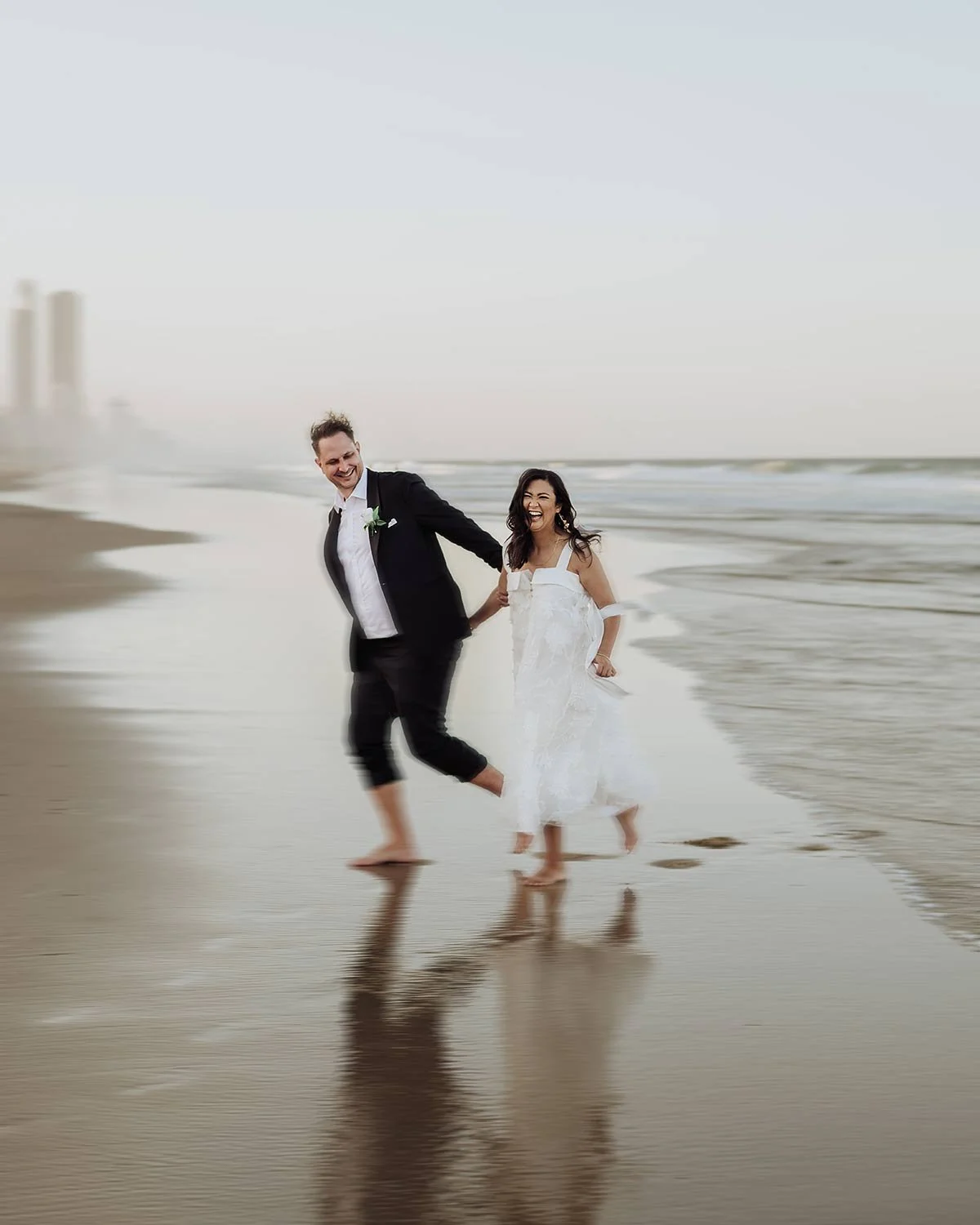A newlywed couple is walking barefoot along a Gold Coast beach, holding hands and laughing as part of a wedding day photoshoot.