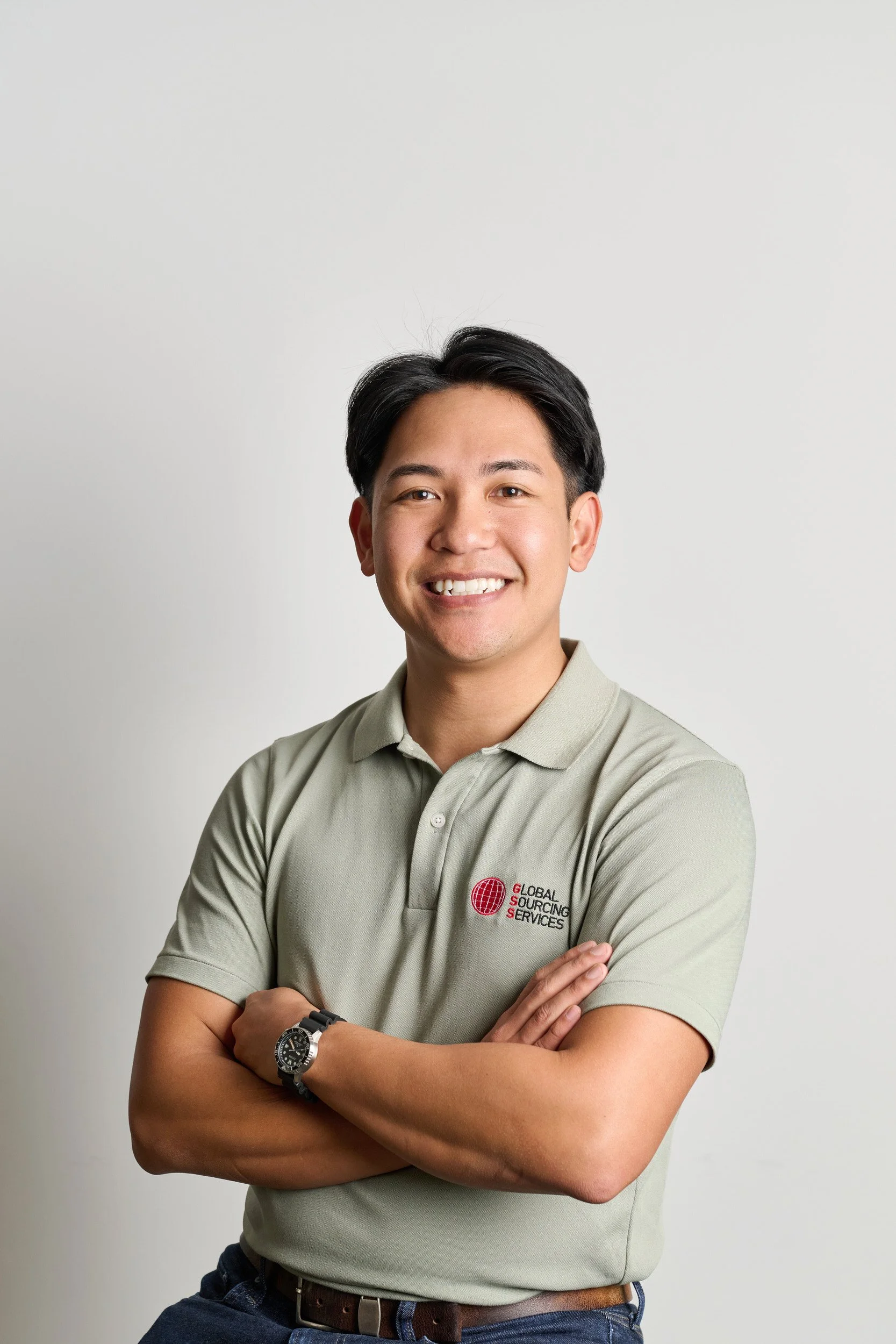 Headshot photography of a young man smiling with arms crossed, wearing a beige polo shirt with a logo that reads 'Global Sourcing Services', a black watch, and dark jeans, professional lit standing against a plain light grey background.