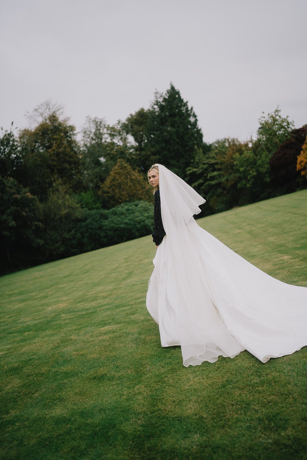 Bride wearing black jumper and full wedding dress with two tier veil looks over her shoulder in large landscaped garden