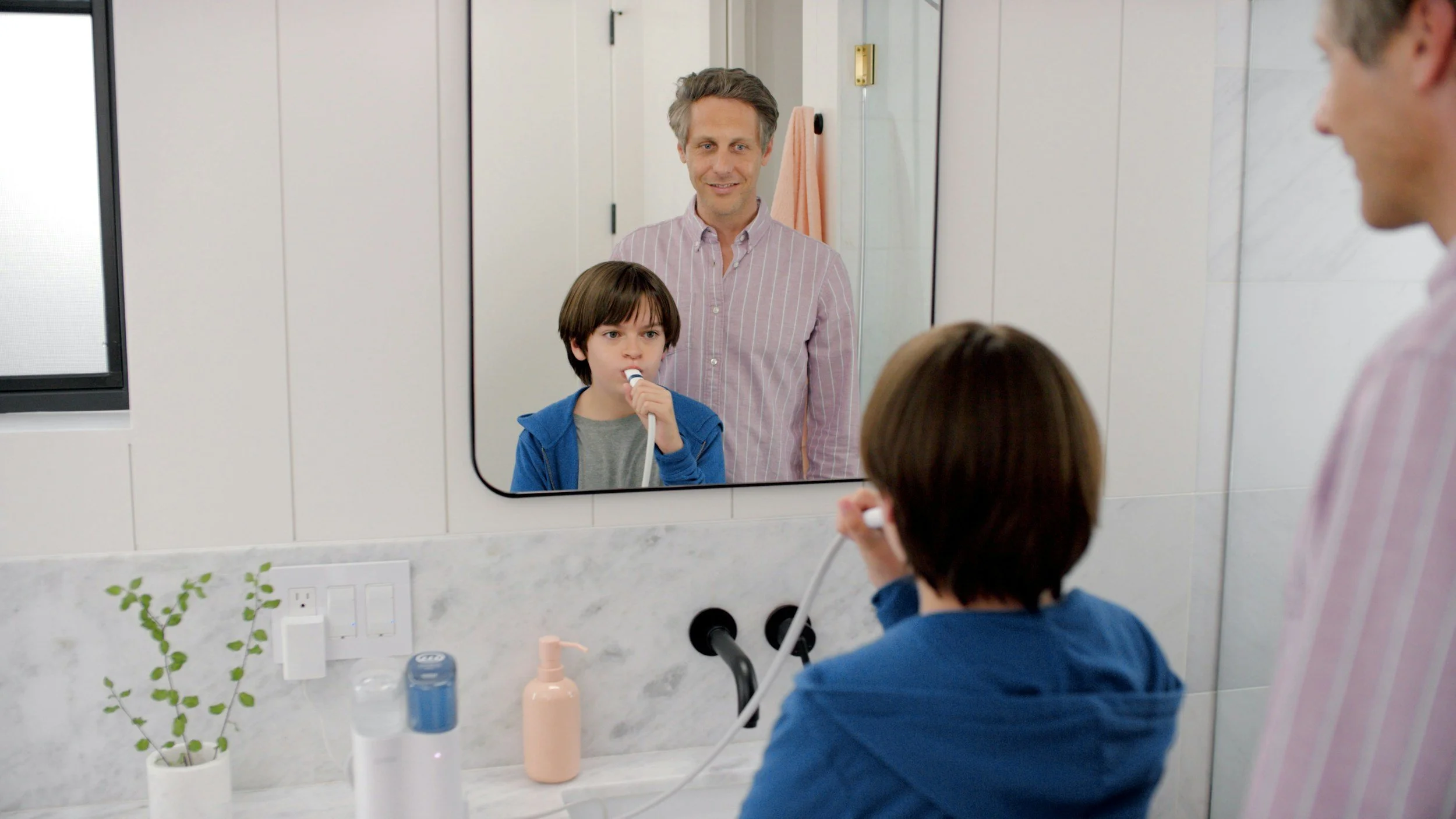 A man with short gray hair stands behind a young boy who is brusing his teeth at the bathroom sink.