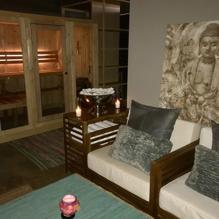 A wood-panelled sauna cabin with lit benches beside a lounge corner of white-cushioned armchairs, candles, rolled towels in a silver bowl and a large Buddha print on the wall