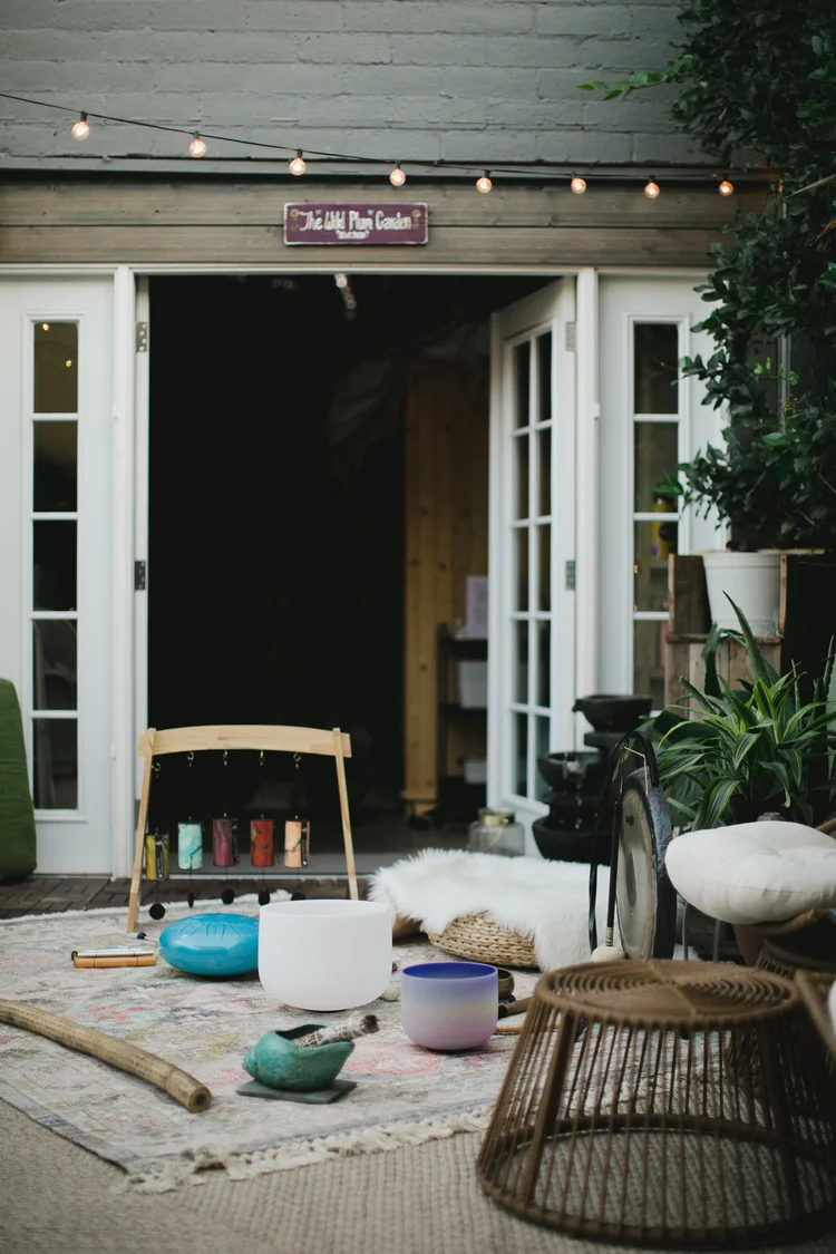 Open French doors under a sign reading The Wild Plum Garden, string lights overhead, with crystal singing bowls, a tongue drum, a gong and a frame of hanging chimes set out on a rug in front