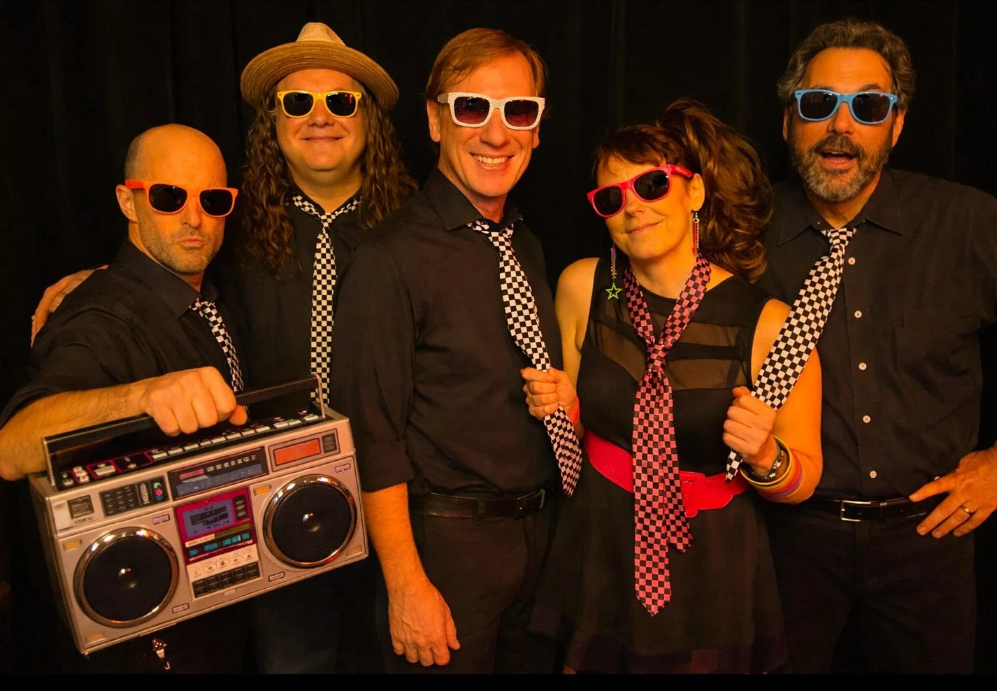 A group of six people dressed in black with checkered accessories, wearing colorful sunglasses, standing together. One person holds a large retro boombox.