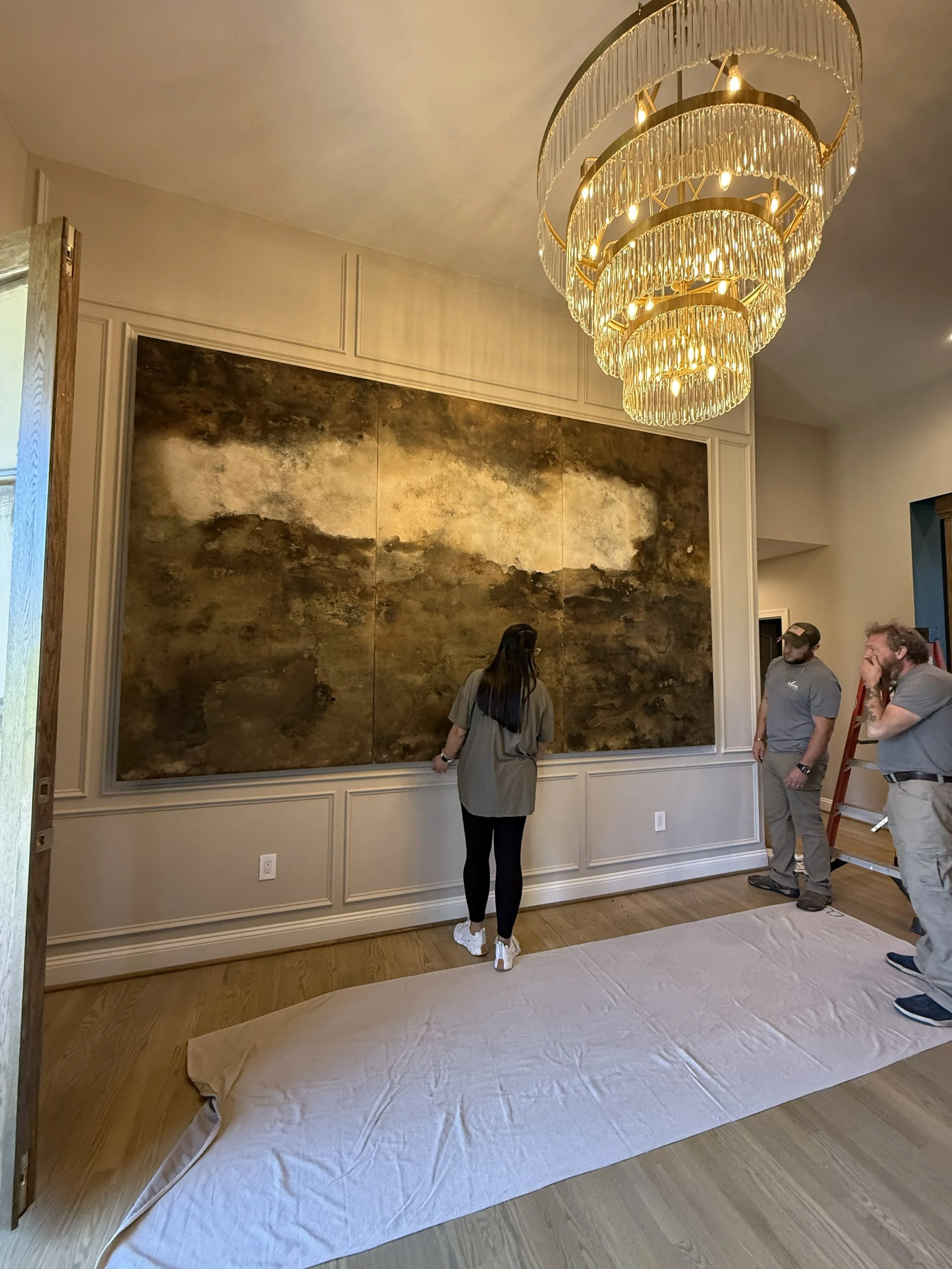 A group of people, including a young woman and two men, are standing in a room with a large, dark, textured painting or wall art behind them. There is a grand chandelier hanging from the ceiling, and a beige sheet or covering on the floor, suggesting a setting for an art exhibit or installation.