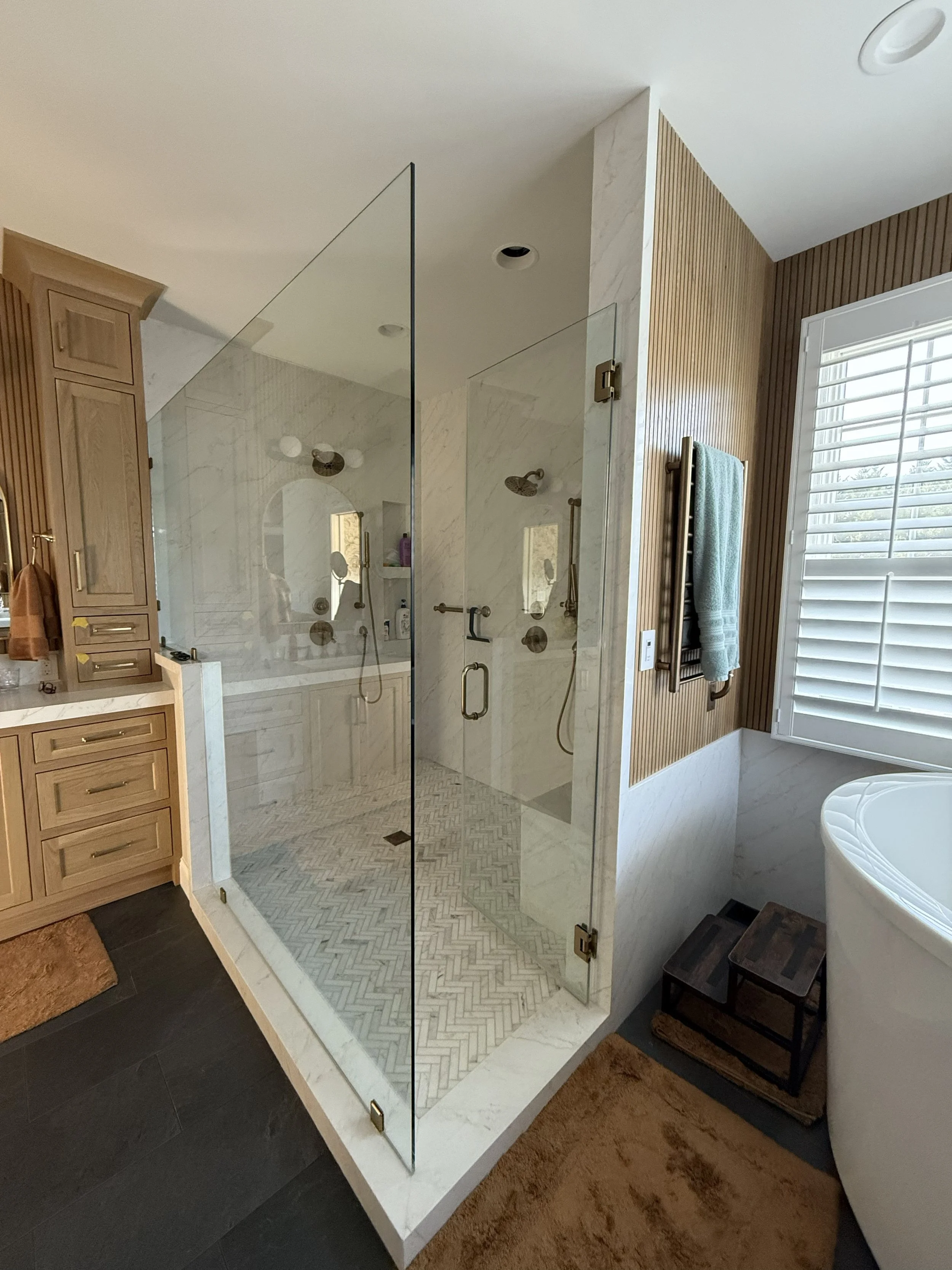 A bathroom with a walk-in shower enclosed by glass, featuring marble walls, multiple showerheads, and a built-in shelf. The bathroom has wooden cabinetry, a towel rack with a towel, a window with shutters, and dark floor tiles with a brown rug.