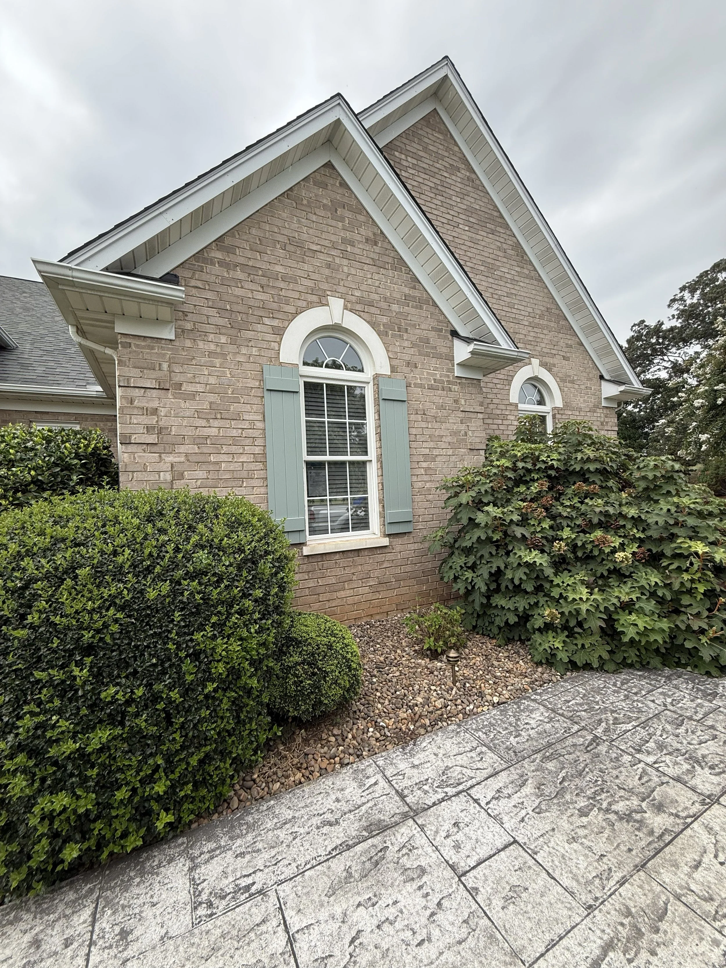 A brick house with white and light blue window shutters, surrounded by bushes and plants, with a stone patio and cloudy sky overhead.