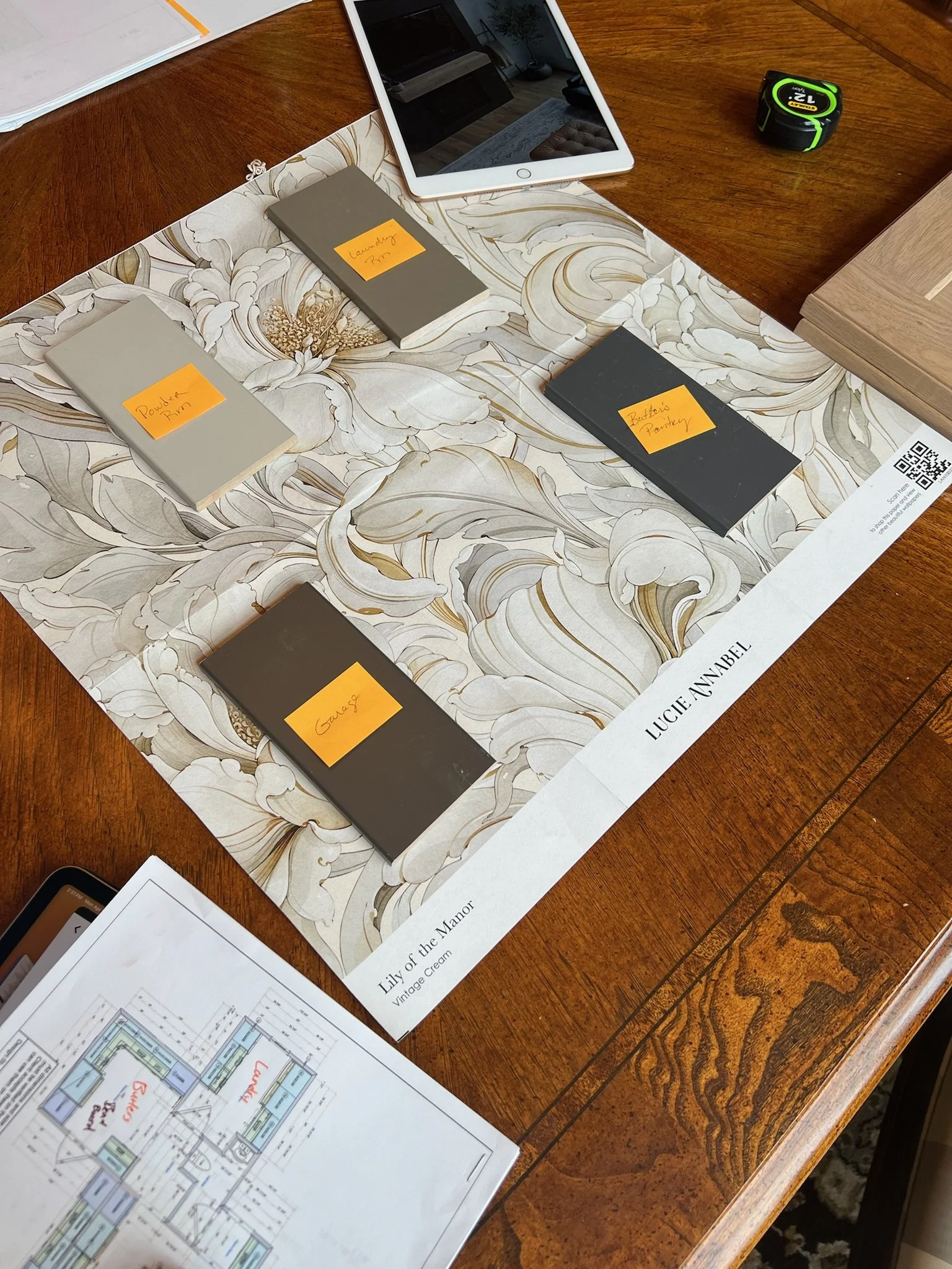 A wooden table with architectural plans, paint swatches, and samples, including a floral patterned paper, with sticky notes labeling the colors such as 'Powder RM', 'Laundry RM', 'Butters Pantery', and 'Gauze'.