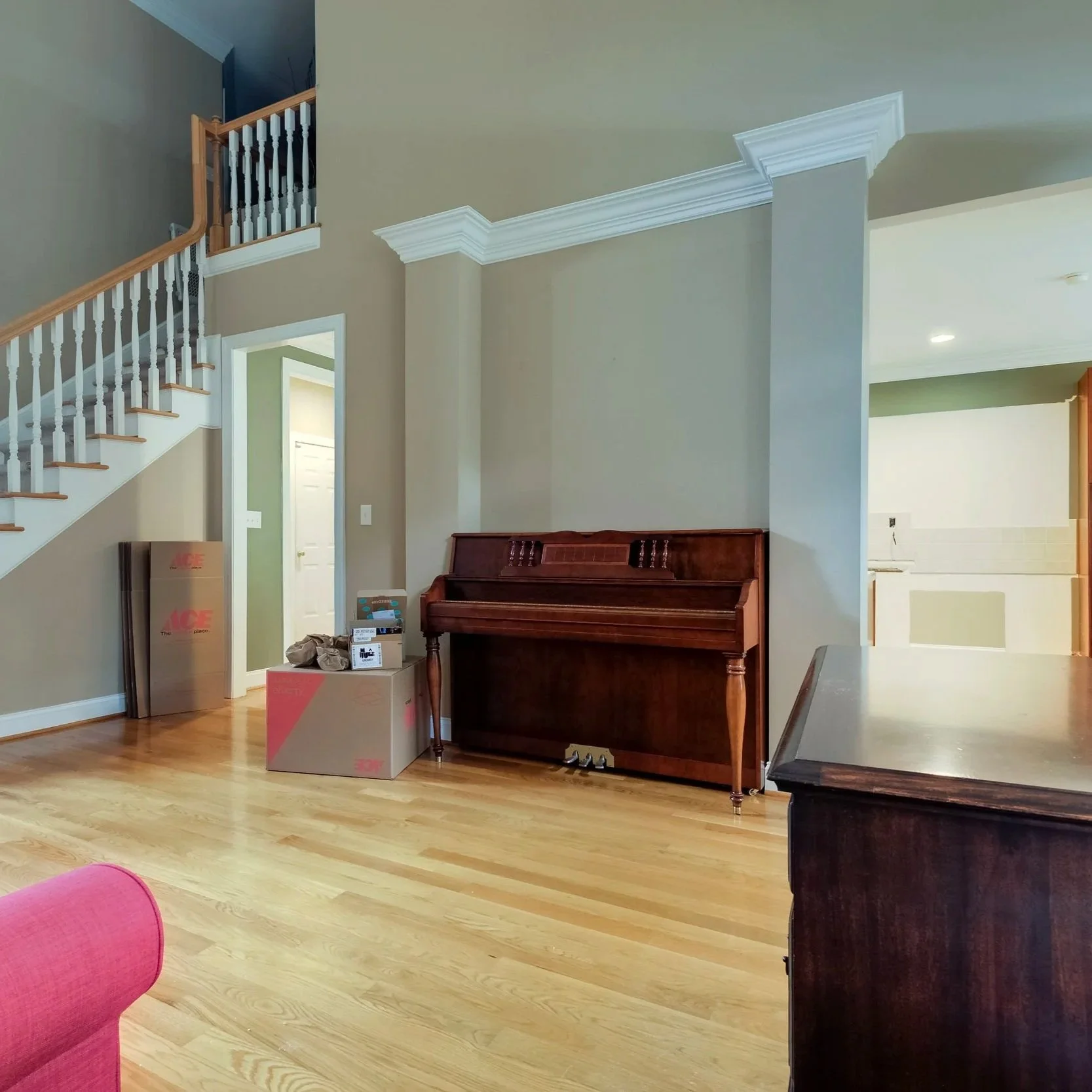 Interior of a home with hardwood floor, beige walls, white crown molding, a staircase with wooden handrail and white balusters, open doorway, and a dark wooden piano. There are cardboard boxes and a pink chair in the foreground.