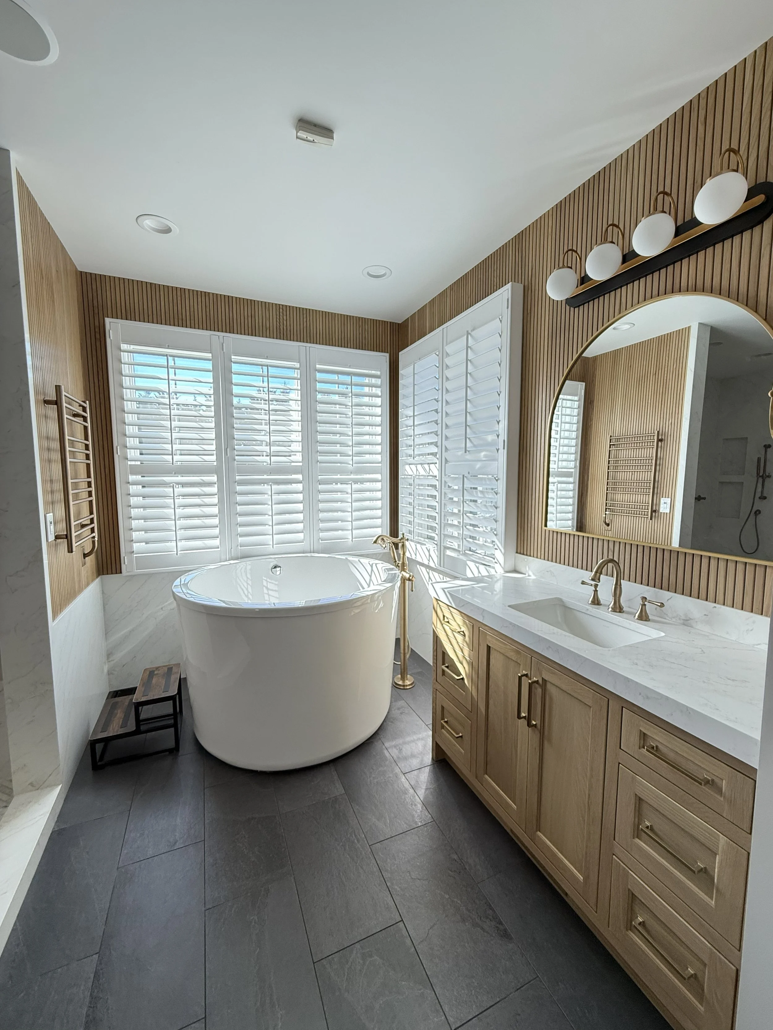 Modern bathroom with a freestanding tub near large window with white shutters, wooden cabinetry with marble countertop, large mirror, and a wall-mounted light fixture.