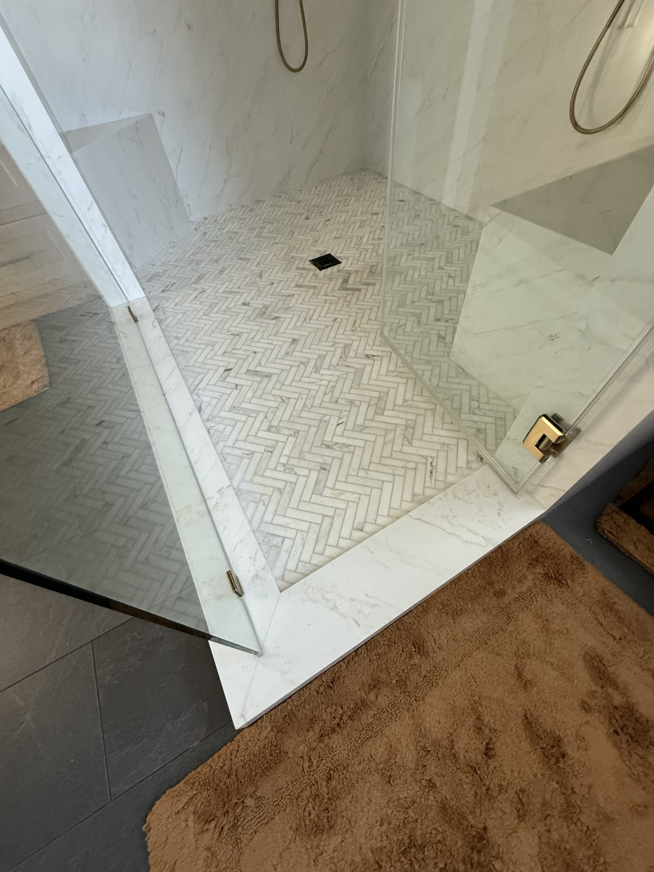 A walk-in shower with marble walls and a herringbone patterned marble floor, a glass door, a black drain, and a handheld showerhead.