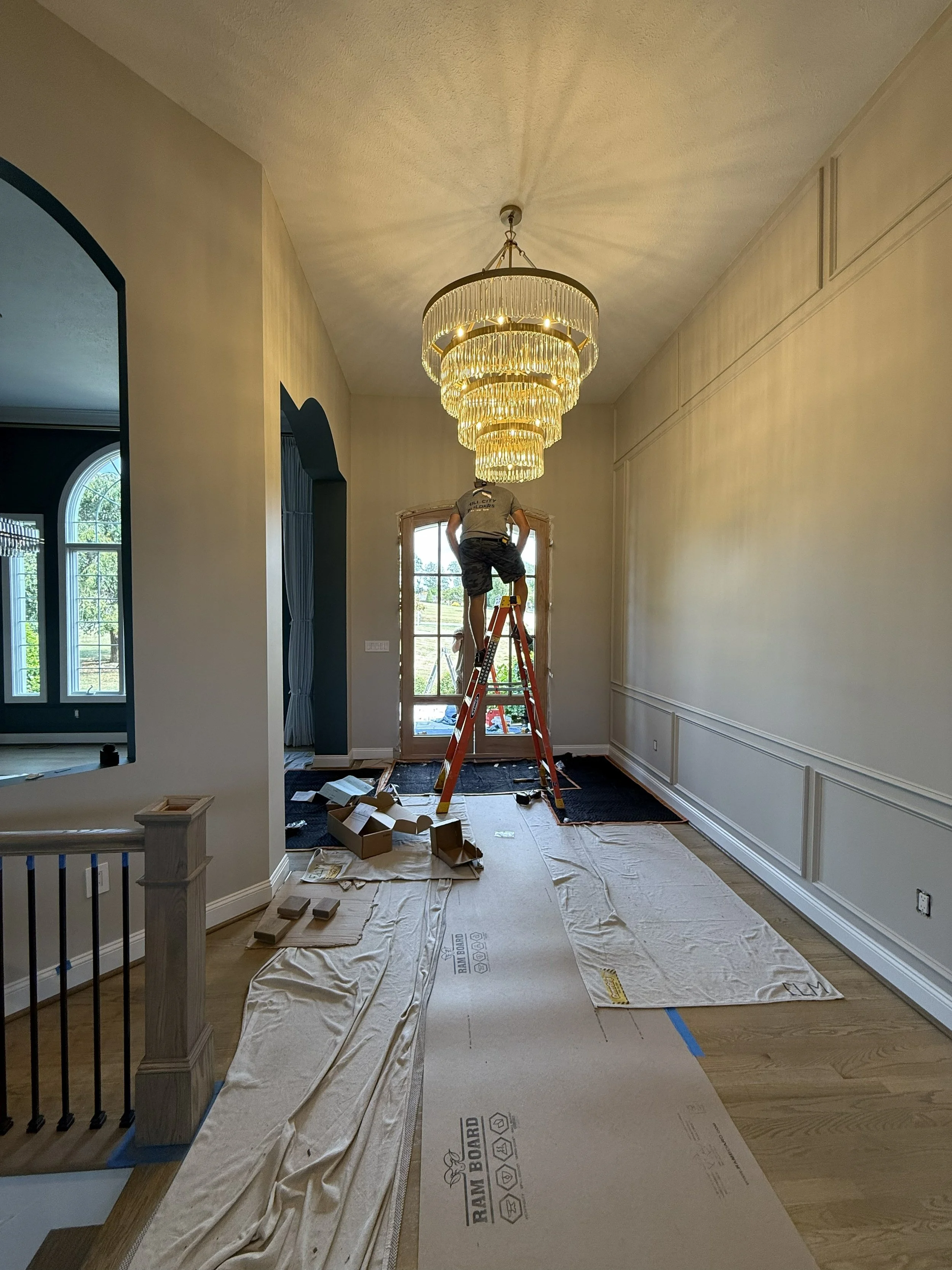 A person standing on a ladder installing or inspecting a large tiered chandelier in a well-lit room with large windows and decorative wall paneling.