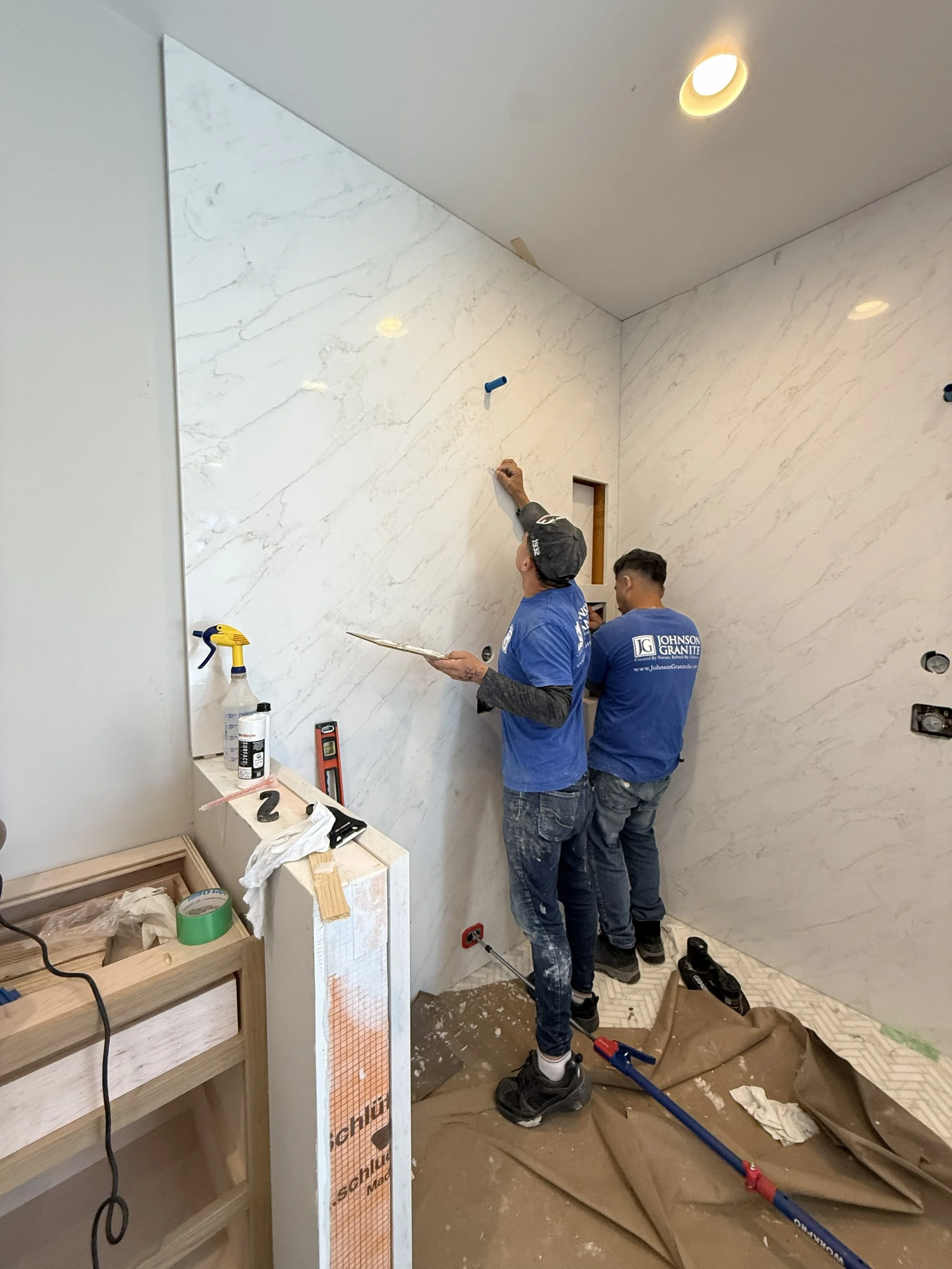 Two workers installing or repairing a marble or stone wall in an indoor setting, with tools and supplies around them.