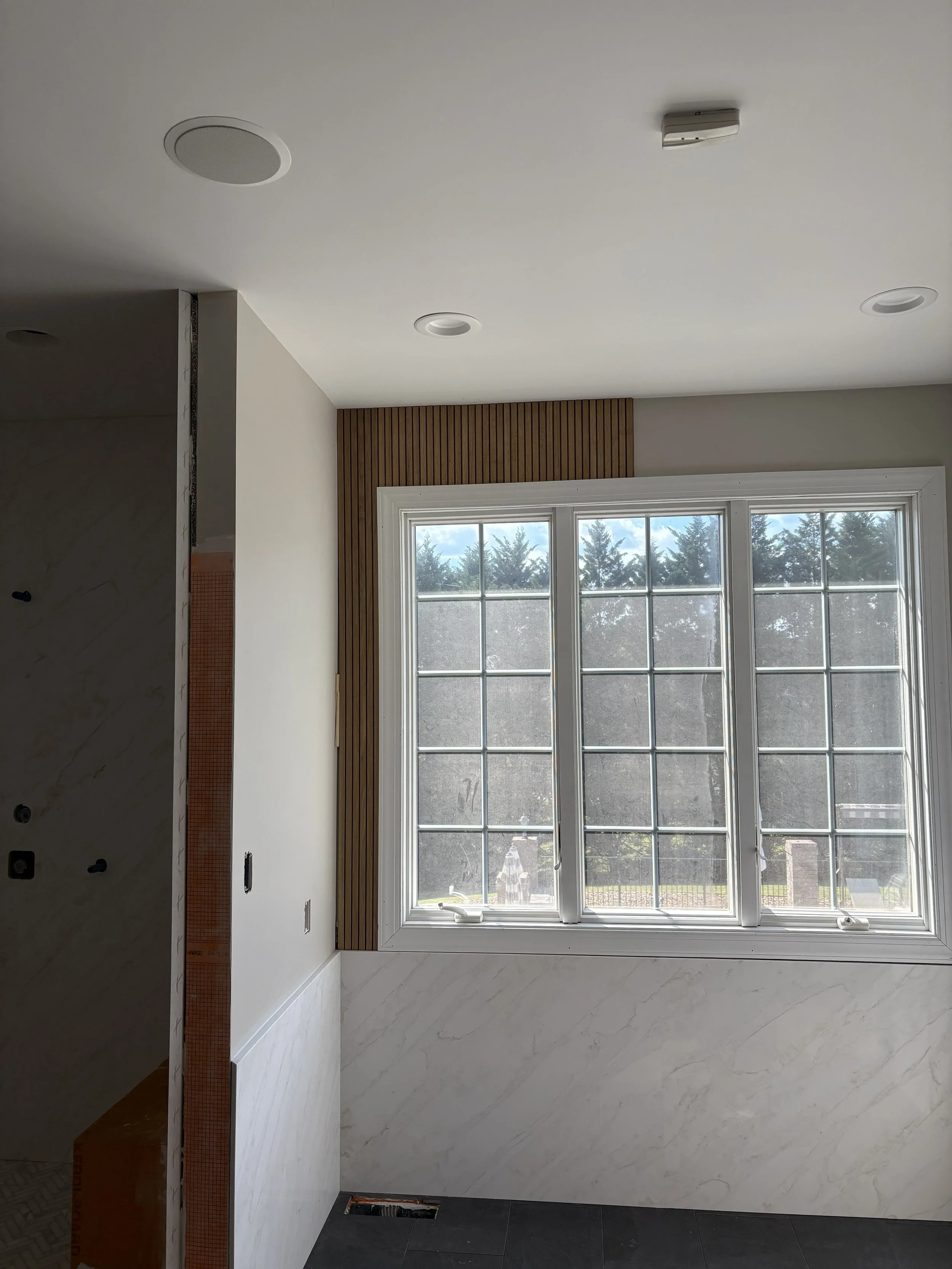 Interior of a room with a large window, white marble walls, and a wooden trim near the ceiling, with some construction or renovation work ongoing.