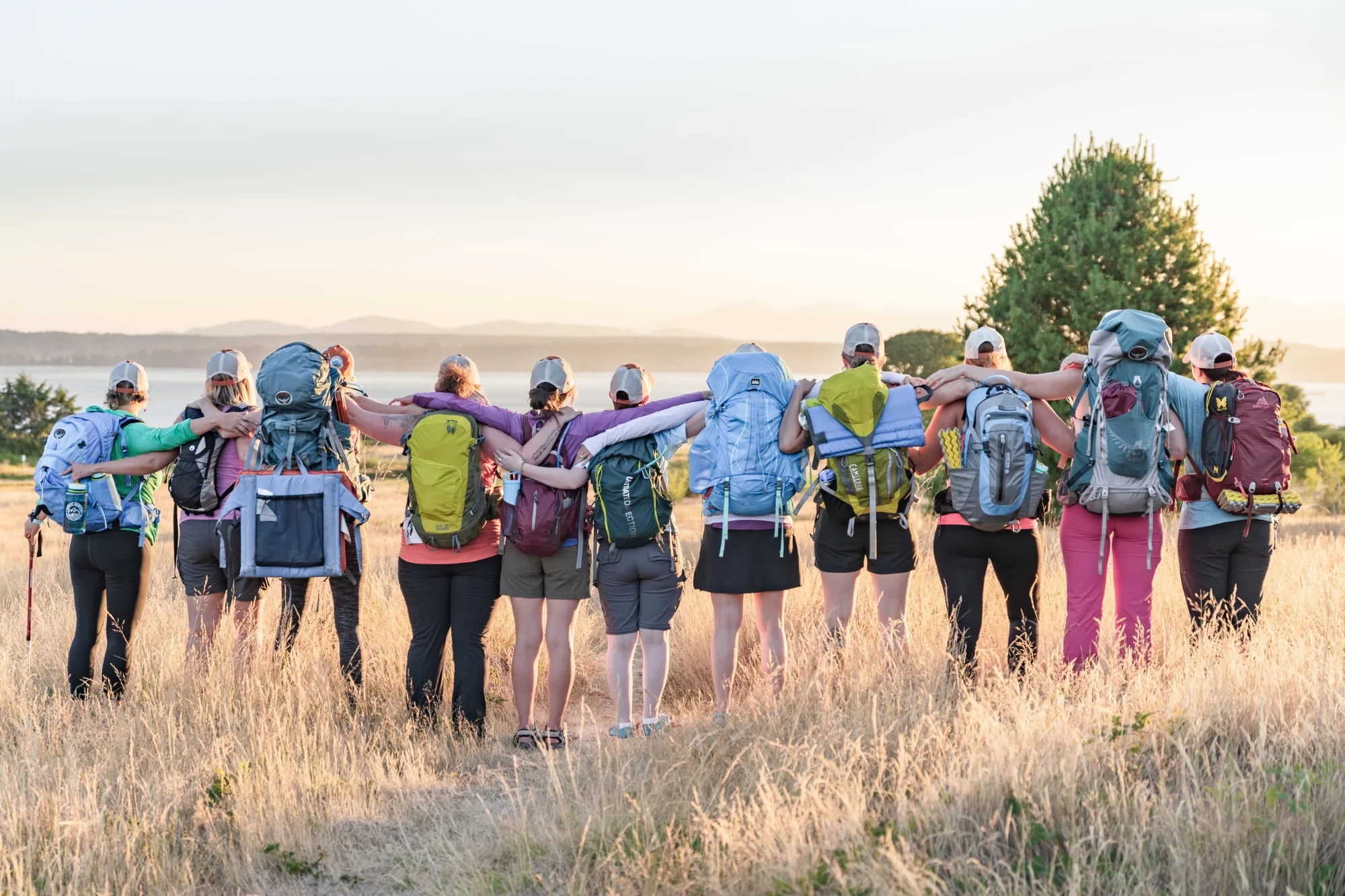 Eleven women with backpacks on facing away from the camera in a field of golden grasses.