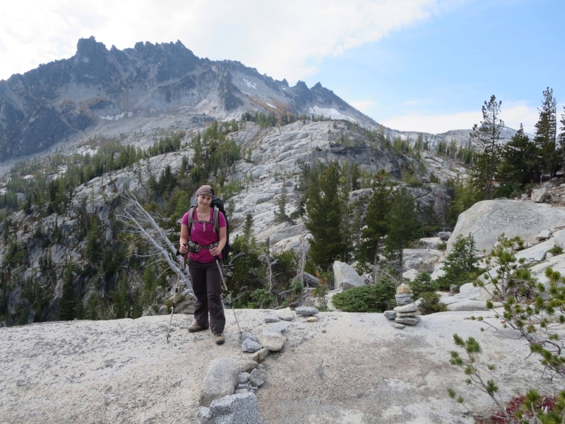 A woman in a pink shirt standing on gray rocks with mountains and trees in the background.