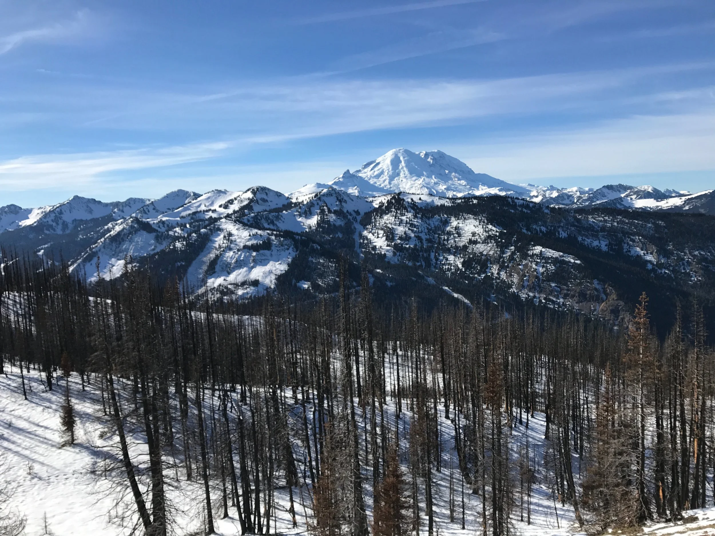 Two hikers on a wilderness trail with patchy snow on the ground. A mountain lake and tree covered mountains in the distance.