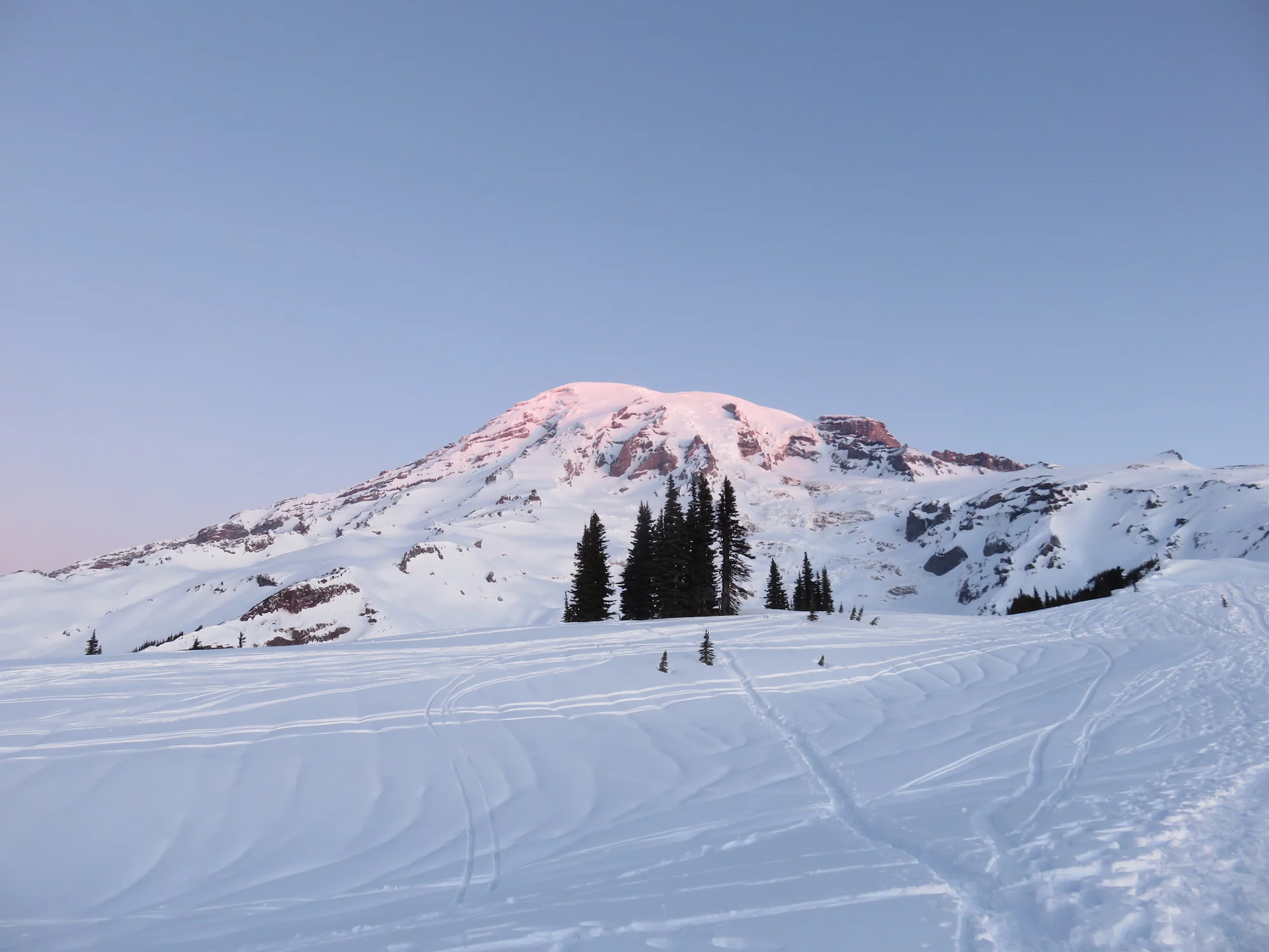 Sunrise light glows pink on the top of a snow covered mountain.