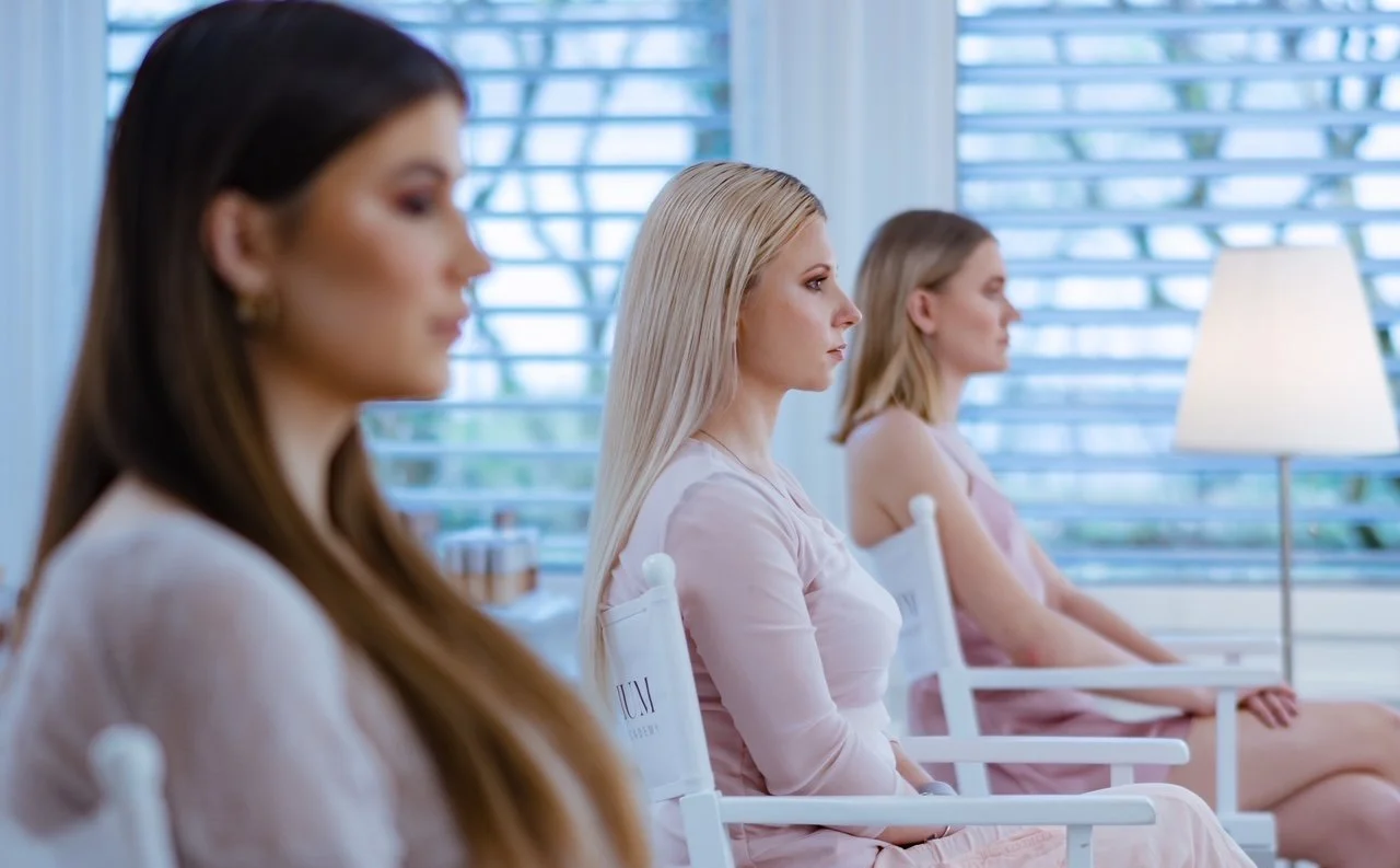 Nahaufnahme eines jungen Erwachsenen mit blonder Haar, auffälligem orangefarbenem Augenschmuck und dezenten Lippenstift, die leicht die Hand an das Kinn legt.
