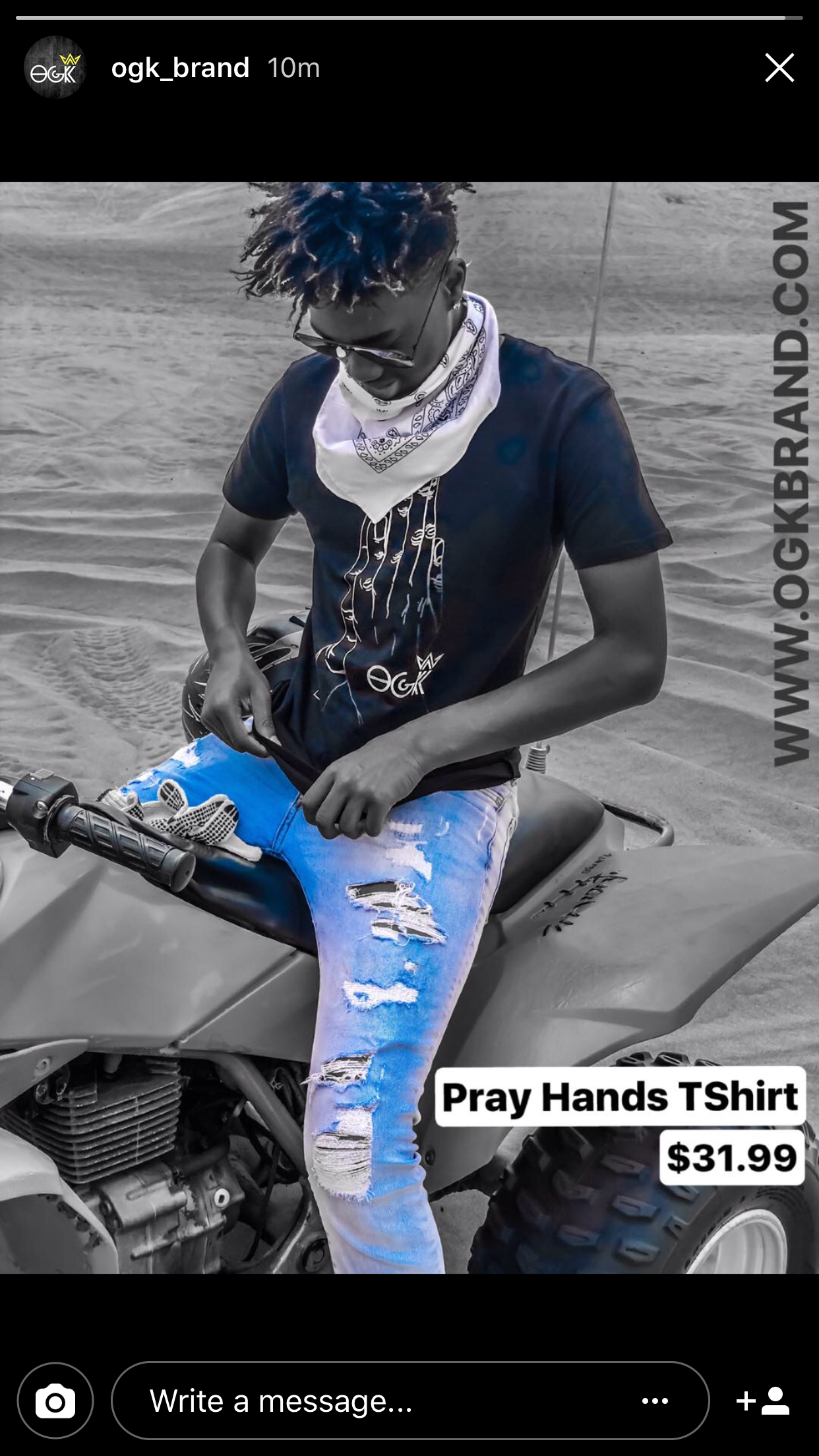 Young man wearing a white bandana around his neck, black sunglasses, a black Pray Hands T-shirt, and ripped blue jeans, sitting on a dirt bike in a desert environment.