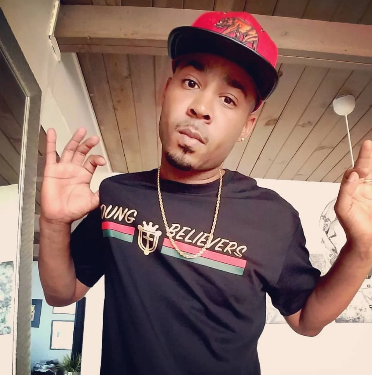 A man wearing a red cap with a lion logo, a black T-shirt with 'YOUNG BELIEVERS' printed on it, a gold chain, and making a hand gesture with both hands, standing indoors with a wooden ceiling.