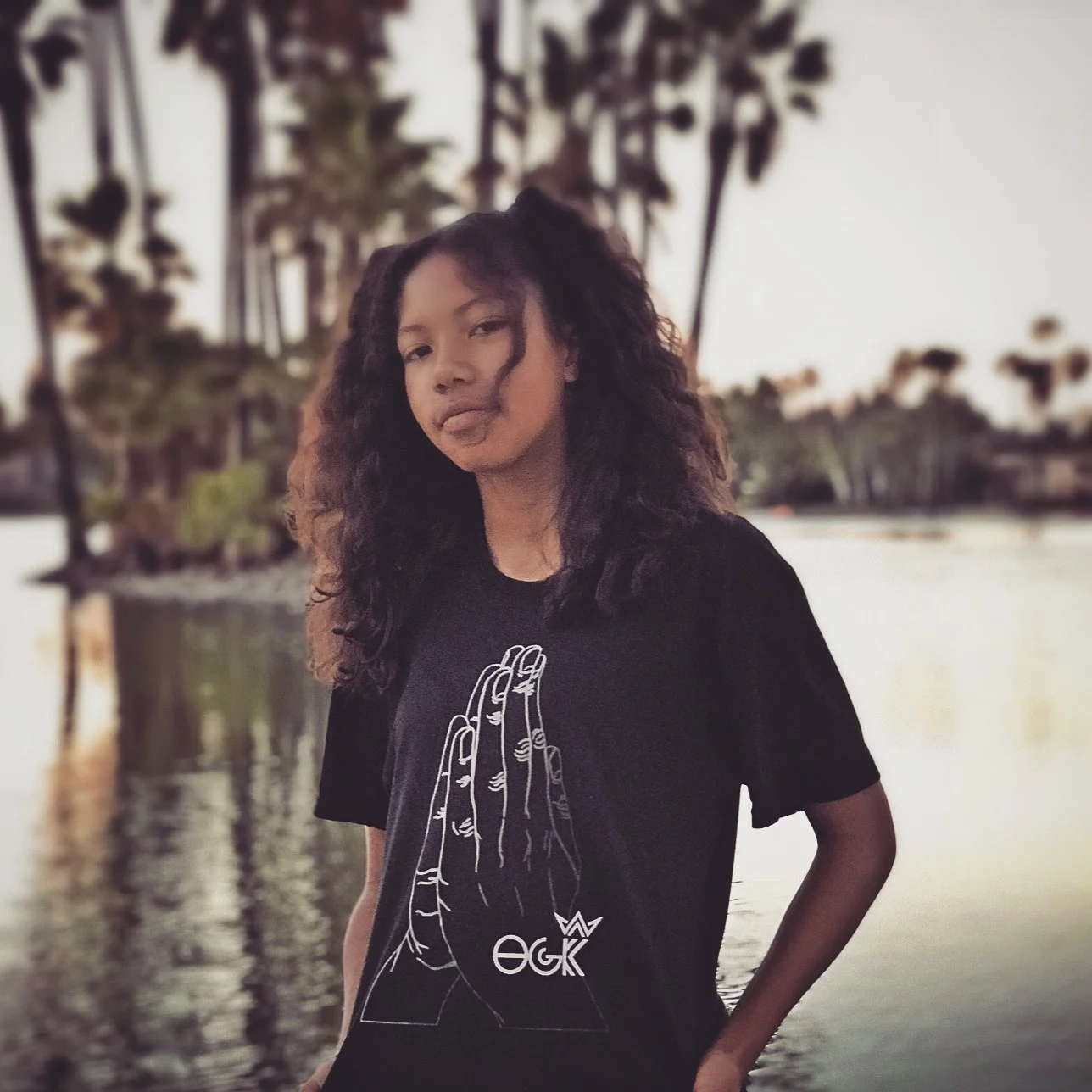 A young woman with curly hair standing near a body of water with trees in the background during sunset.