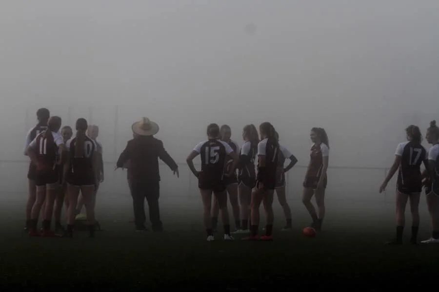 A group of young women soccer players in uniform gathered on a foggy field, listening to with Craig Gunn performance coach leading.