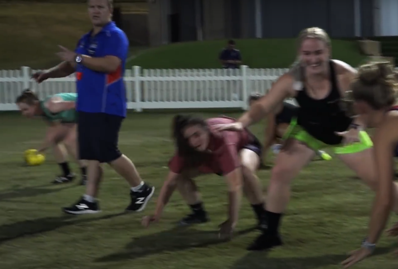 Group of people participating in a AFL or women's Australian football fitness or outdoor exercise session on a grassy field, some in crawling positions, with a white picket fence and a building in the background. Craig Gunn performance coach