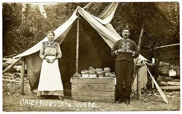 Photo postcard of Mi’kmaq chief Noel and his wife. Between them is a display of their baskets. Circa 1910