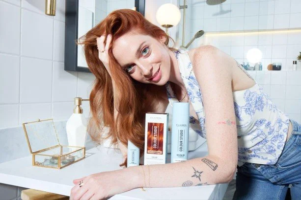 A woman with red hair leaning on a bathroom counter, displaying skincare products and tattoos.
