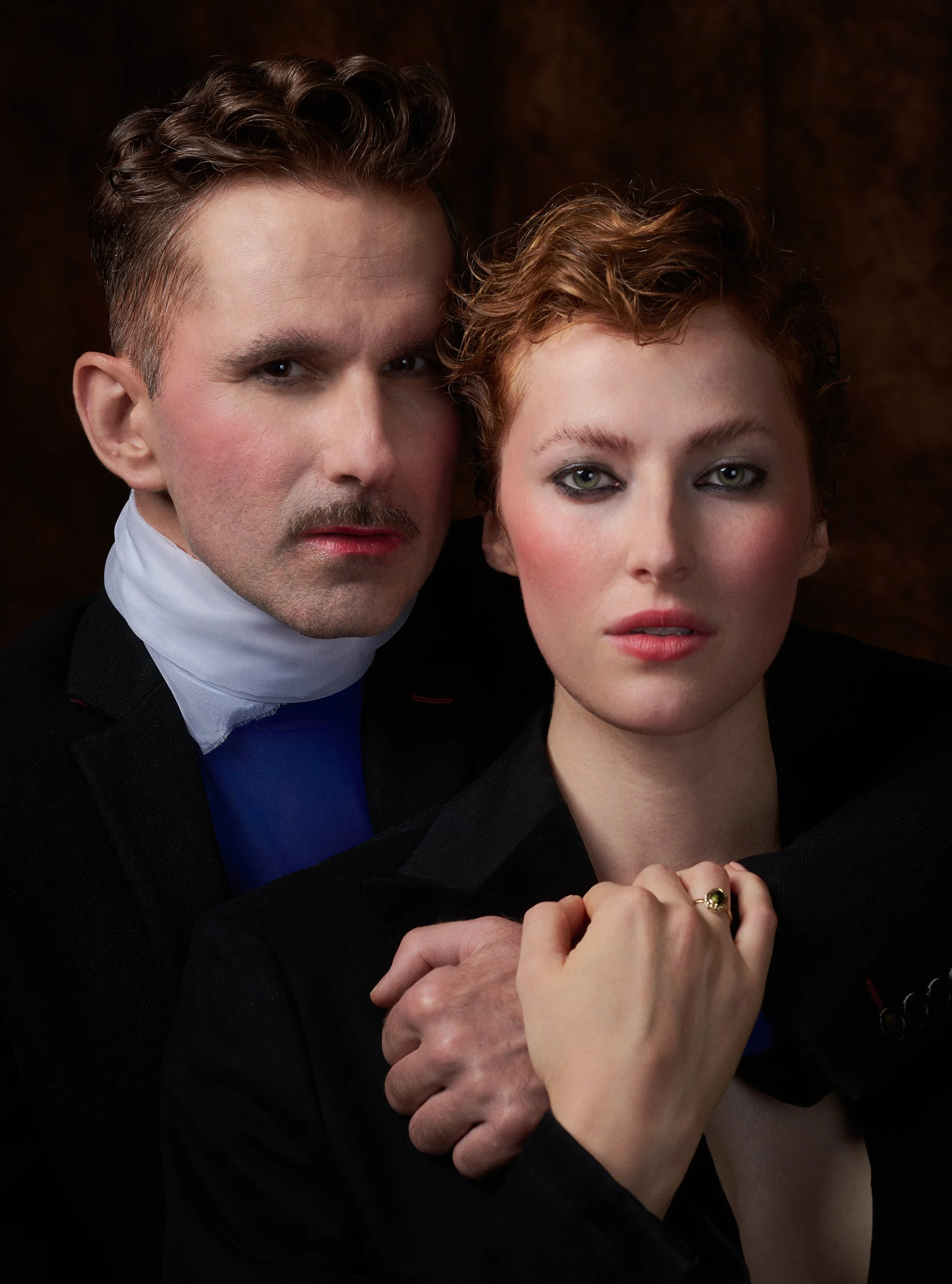 A man with short curly brown hair, gray eyes, and a mustache, wearing a tuxedo, and a woman with short curly red hair and light green eyes, wearing makeup, jewelry, and a black jacket, are close together with their hands clasped.