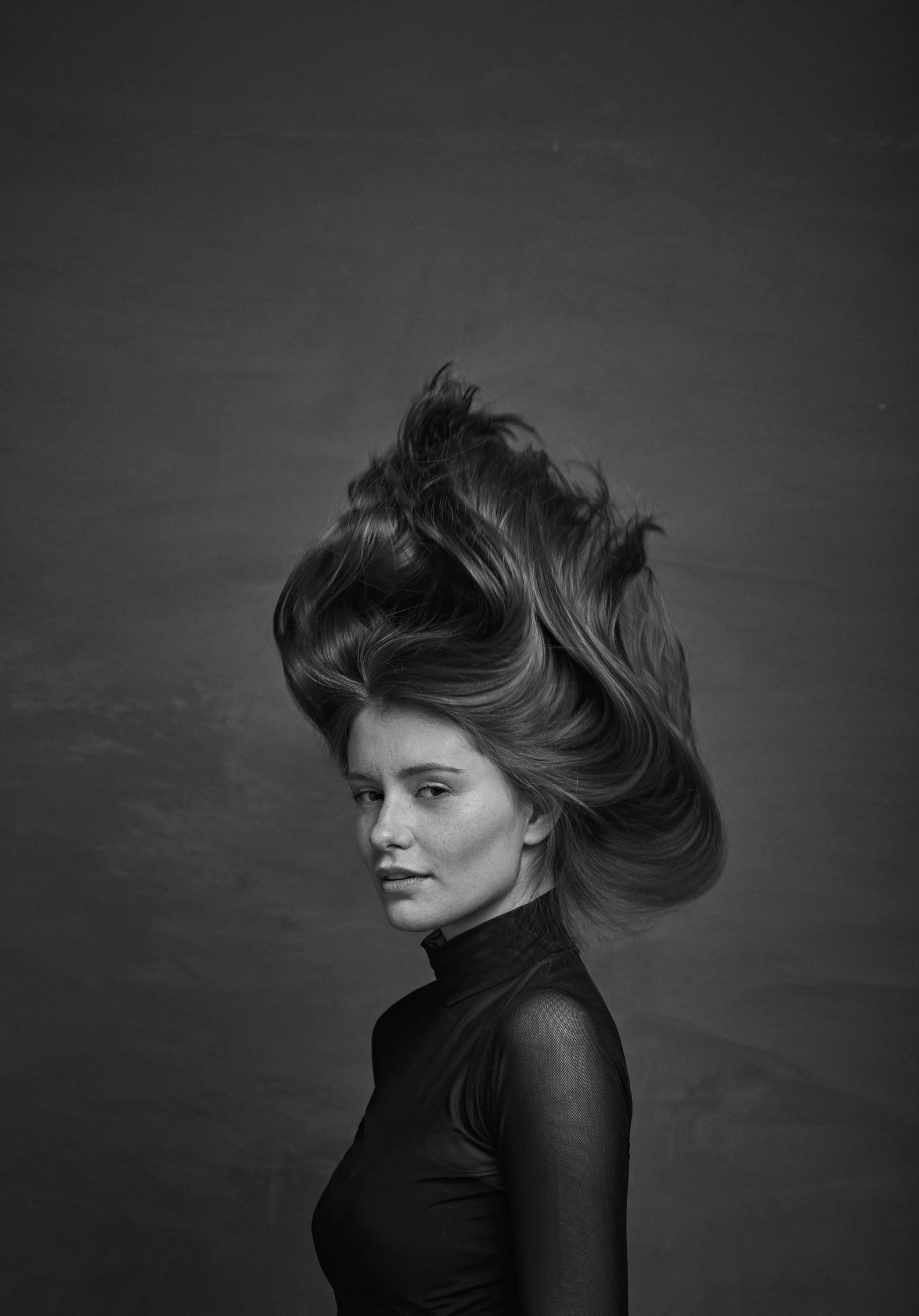 A black and white photo of a woman with wavy hair blowing in the wind, looking over her shoulder, wearing a dark turtleneck and a sheer long sleeve top.