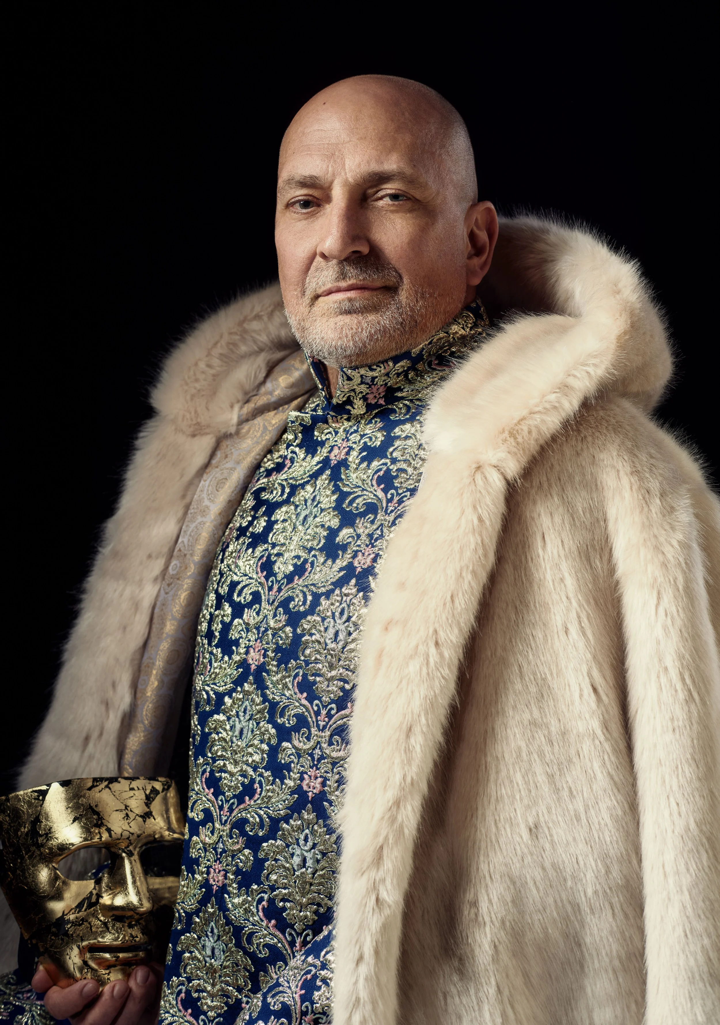 A man with a bald head and gray beard wearing a luxurious white fur coat. He is holding a gold mask with a black and gold design, and he is dressed in an ornate, patterned garment with a brocade design.