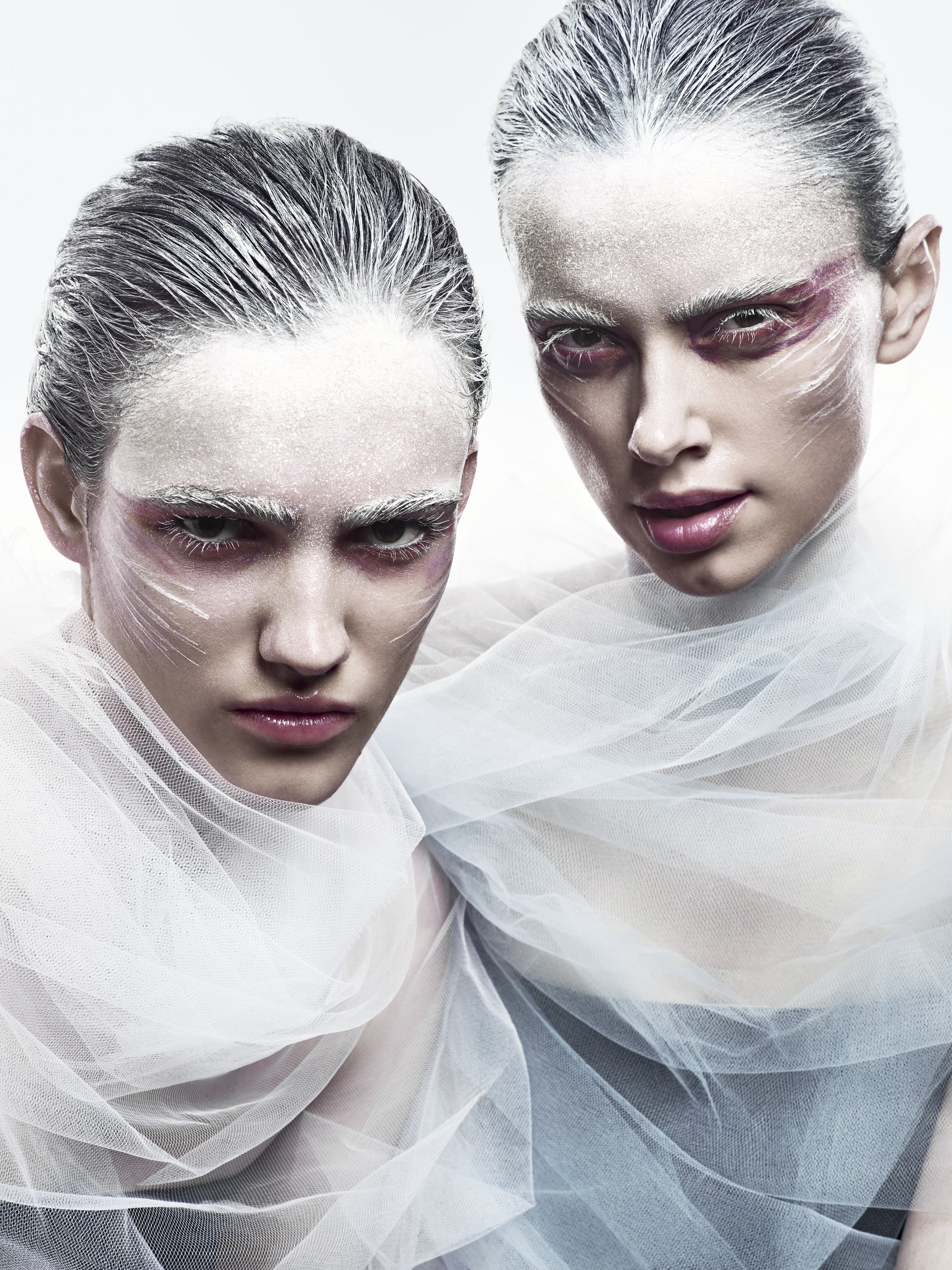 Two women with white makeup and painted eyes, dressed in sheer white fabric, posing closely together against a white background.