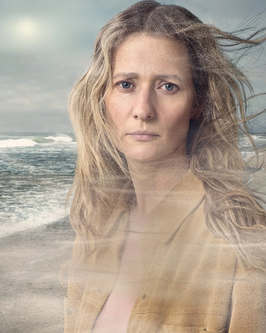 A woman with long tousled hair looking directly at the camera, partially transparent, superimposed over a beach scene with waves and cloudy sky.