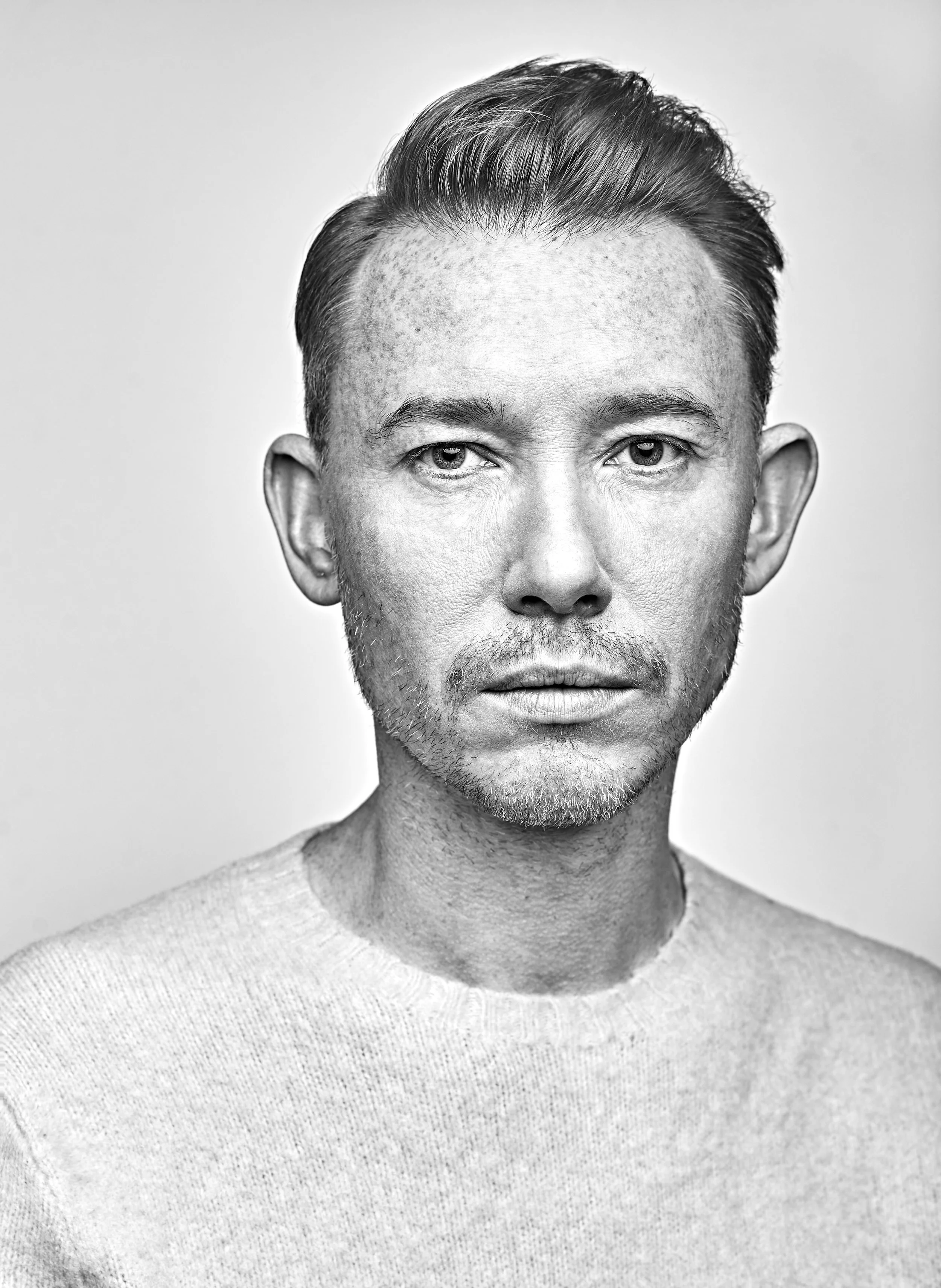 Black and white portrait of a man with short hair, light facial hair, wearing a crew neck sweater, facing the camera with a neutral expression.