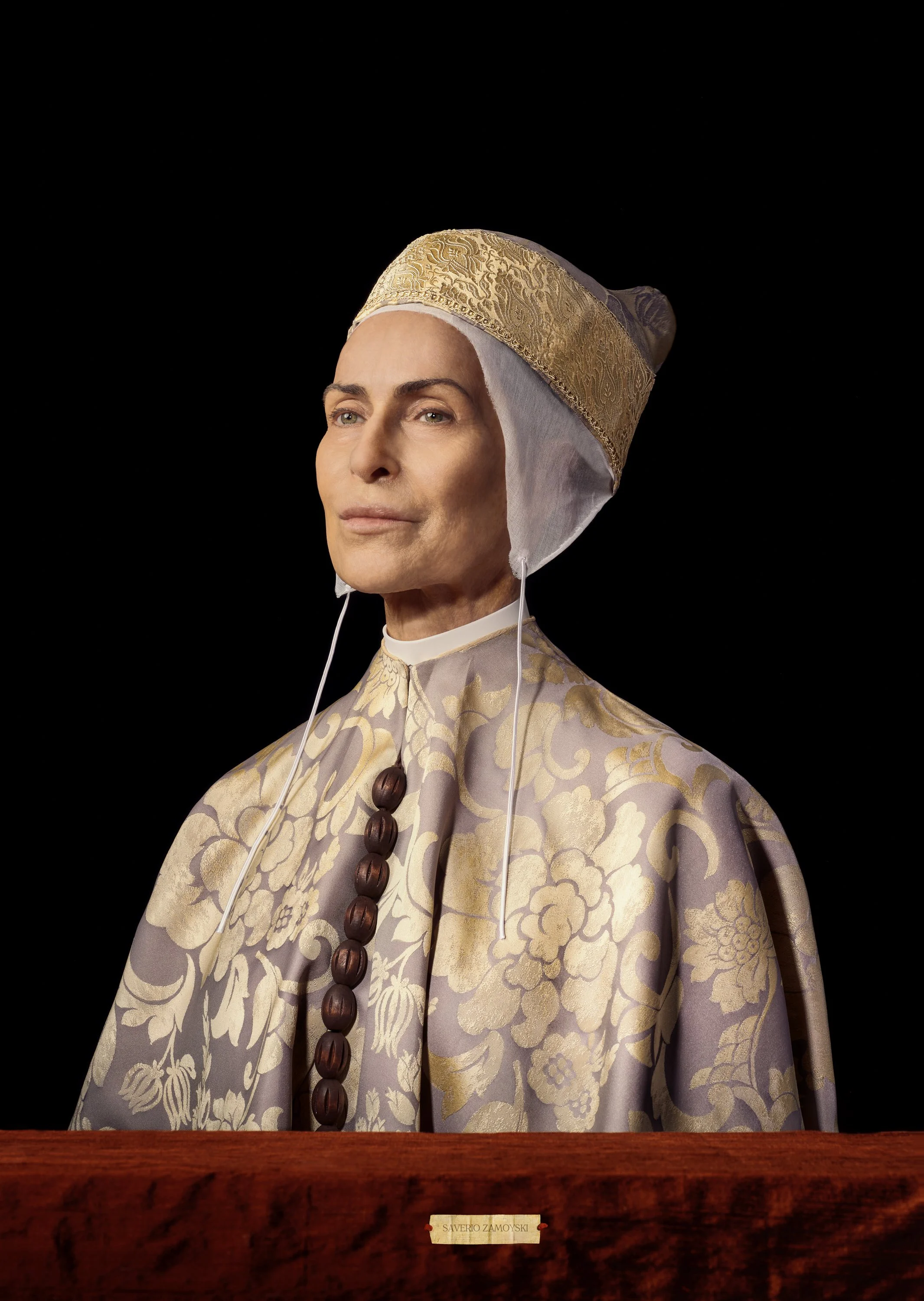 Statue of a woman wearing a traditional headdress and a patterned garment, standing behind a wooden desk with a nameplate that reads 'Saverio Zamoyski'. The stylized portrait of Danuta Stenka