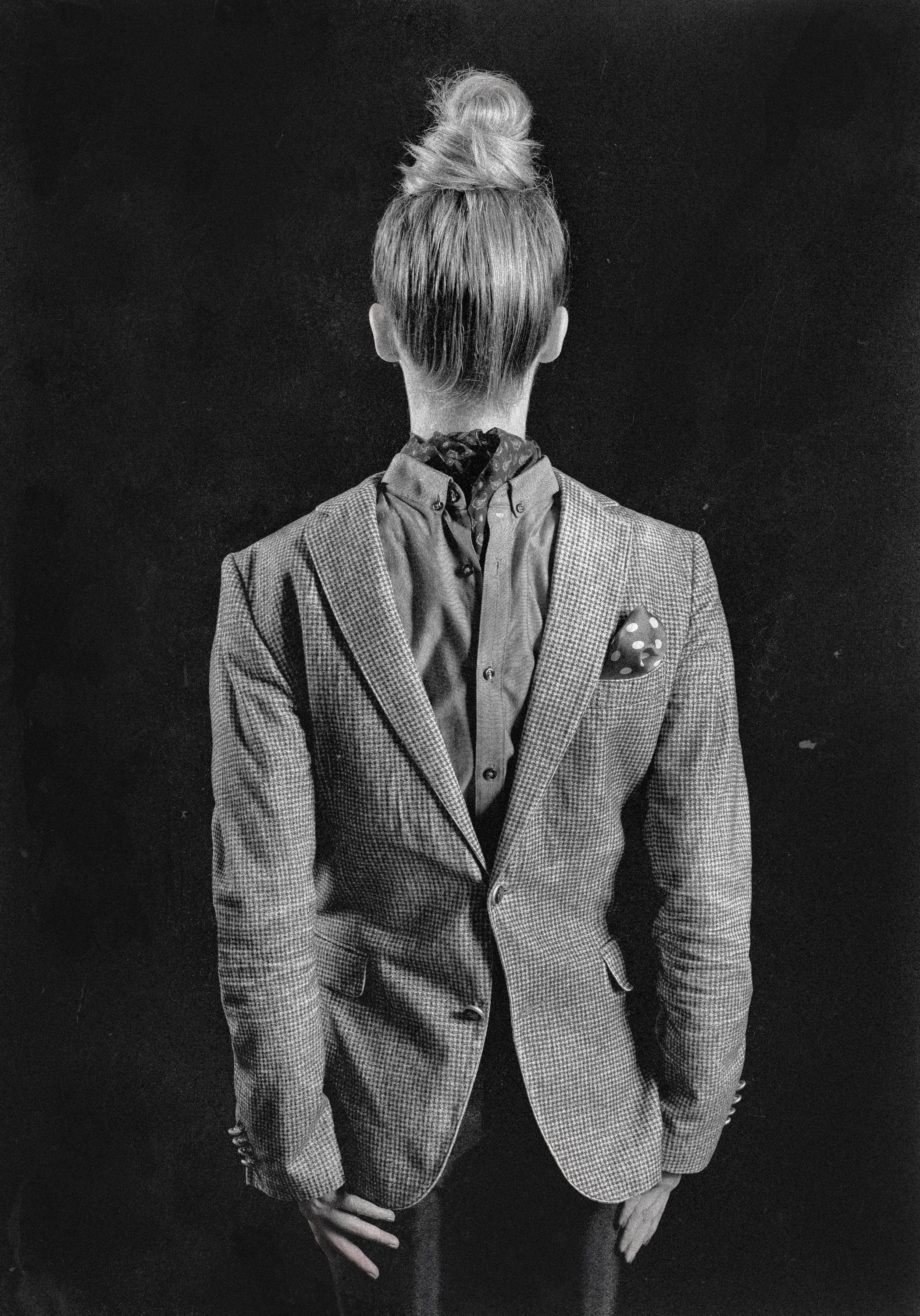 Black and white photograph of a man standing against a dark background, viewed from behind. He is wearing a plaid blazer over a shirt with a patterned scarf and has his hair tied up in a bun.