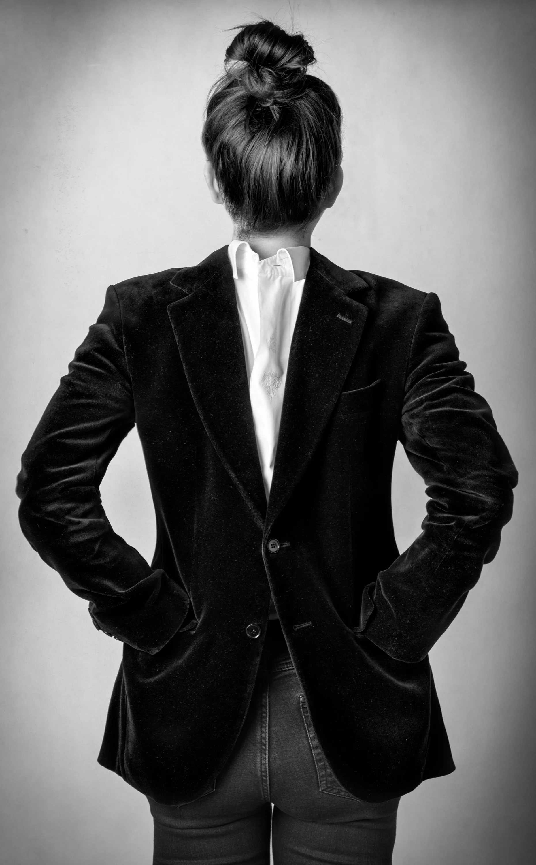 Black and white photo of a person from behind, wearing a blazer and jeans, hair tied in a bun, standing with hands on hips.